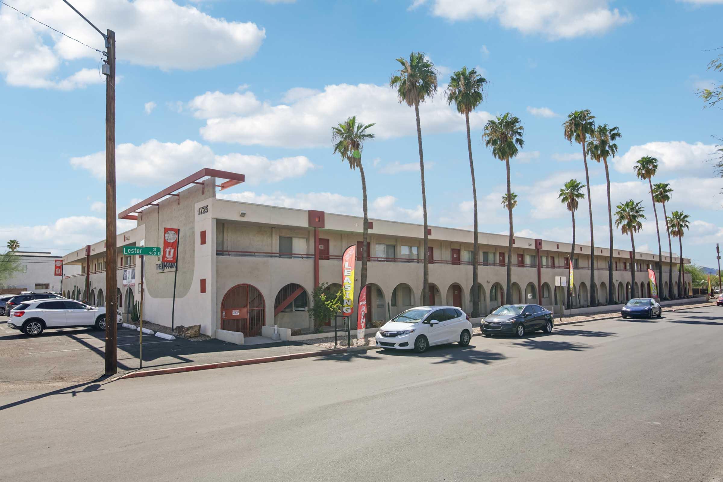 A low-rise motel with a beige exterior and arches, situated along a street lined with palm trees. Several parked cars are visible in the foreground. The sky is partly cloudy, and the setting appears to be in a warm climate. A sign at the entrance indicates the name of the motel.