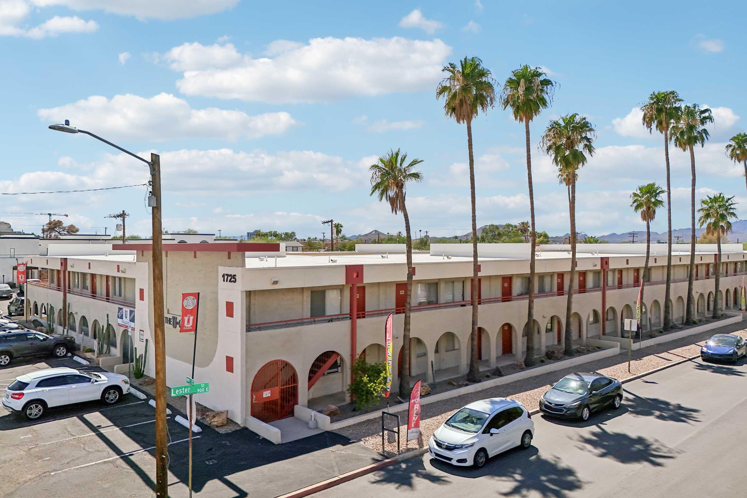 A low-rise building with multiple units featuring palm trees in front and parked cars along the street. The building has a light-colored exterior with red accents and signs. Blue sky with clouds is visible above. The scene depicts a sunny day in an urban area.