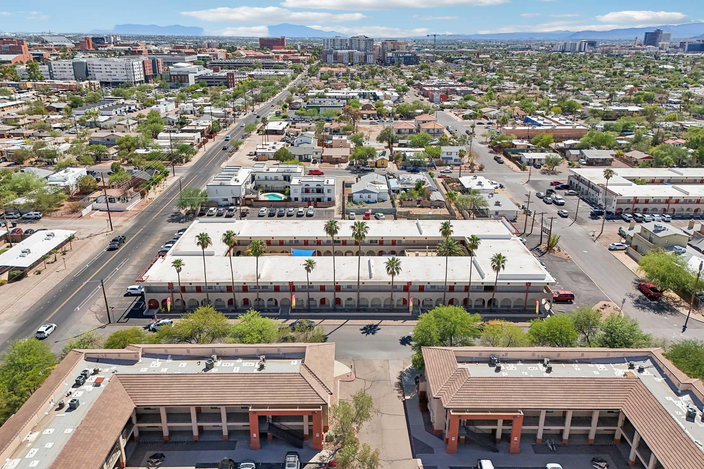 Aerial view of a neighborhood showing a mix of residential and commercial buildings, including low-rise apartments and a motel with a swimming pool. Surrounding areas feature trees, roads, and distant mountains under a partly cloudy sky.
