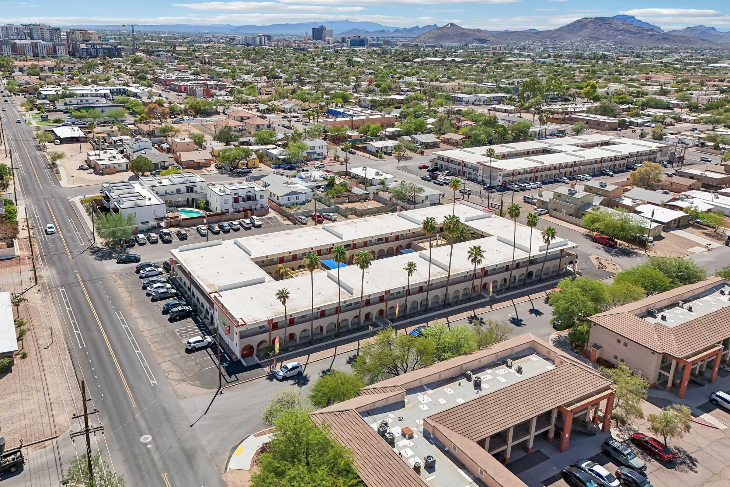 Aerial view of a residential area featuring a low-rise building with multiple parking spaces, palm trees, and roads. In the background, various houses and commercial buildings are visible, along with distant mountains and a clear blue sky.