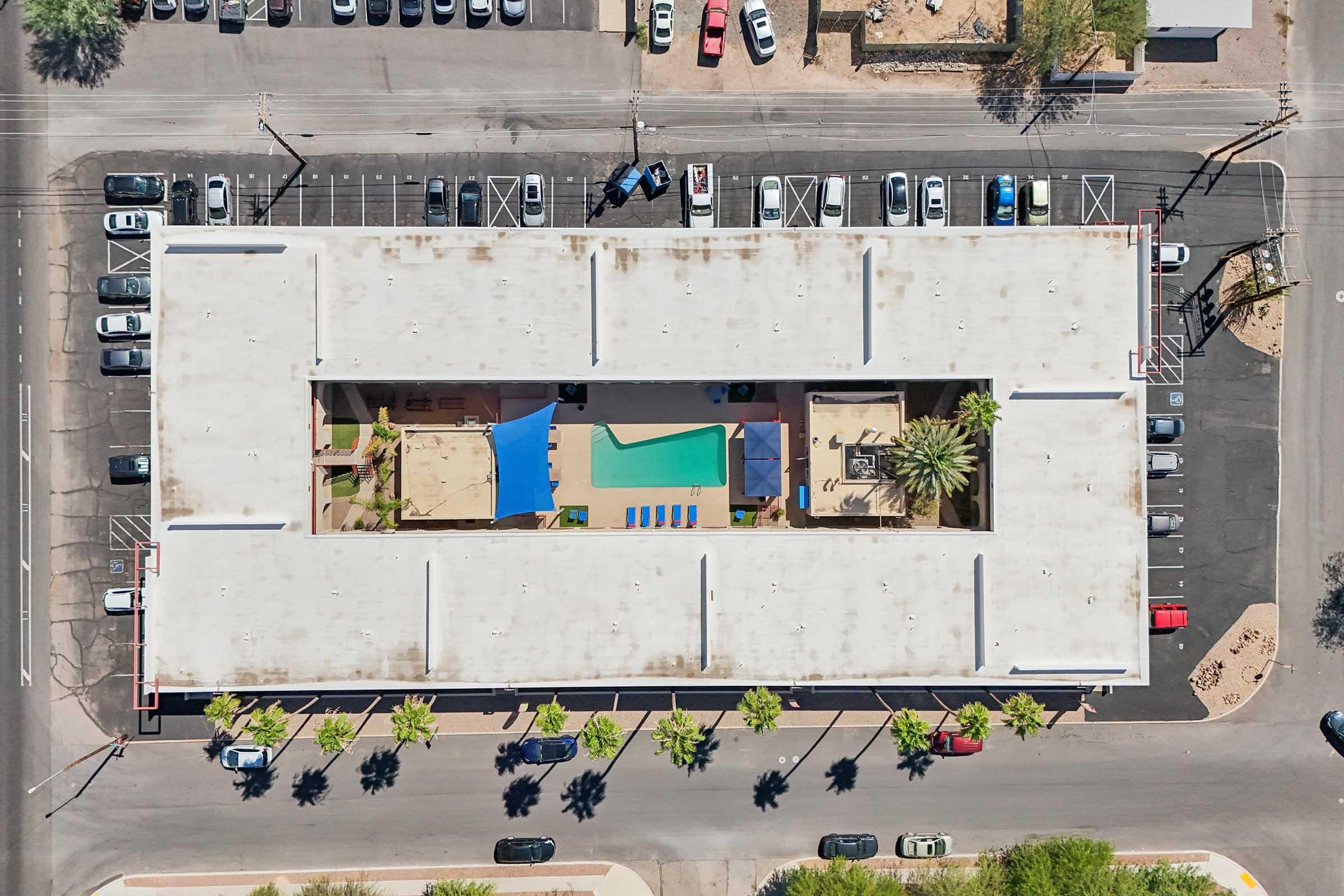 Aerial view of a building complex featuring a central courtyard with a swimming pool surrounded by lounge chairs. Palm trees line the courtyard, and parked cars are visible in the surrounding parking lots. The building has a flat roof and is situated in a suburban area.