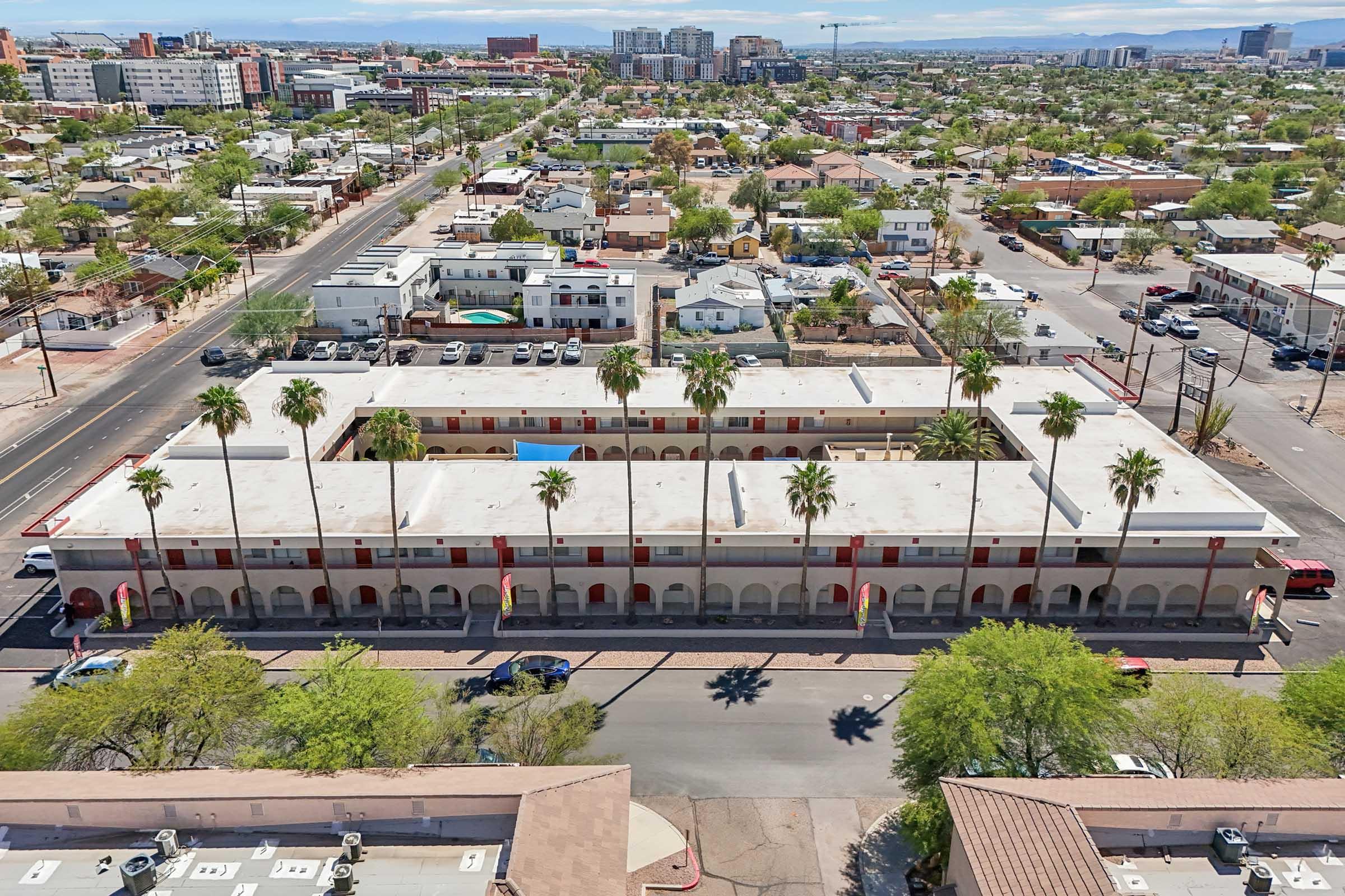 An aerial view of a neighborhood featuring a long, single-story building with red accents and palm trees lining the front. The surrounding area includes various residential structures and a street. In the distance, modern buildings and a clear blue sky are visible.