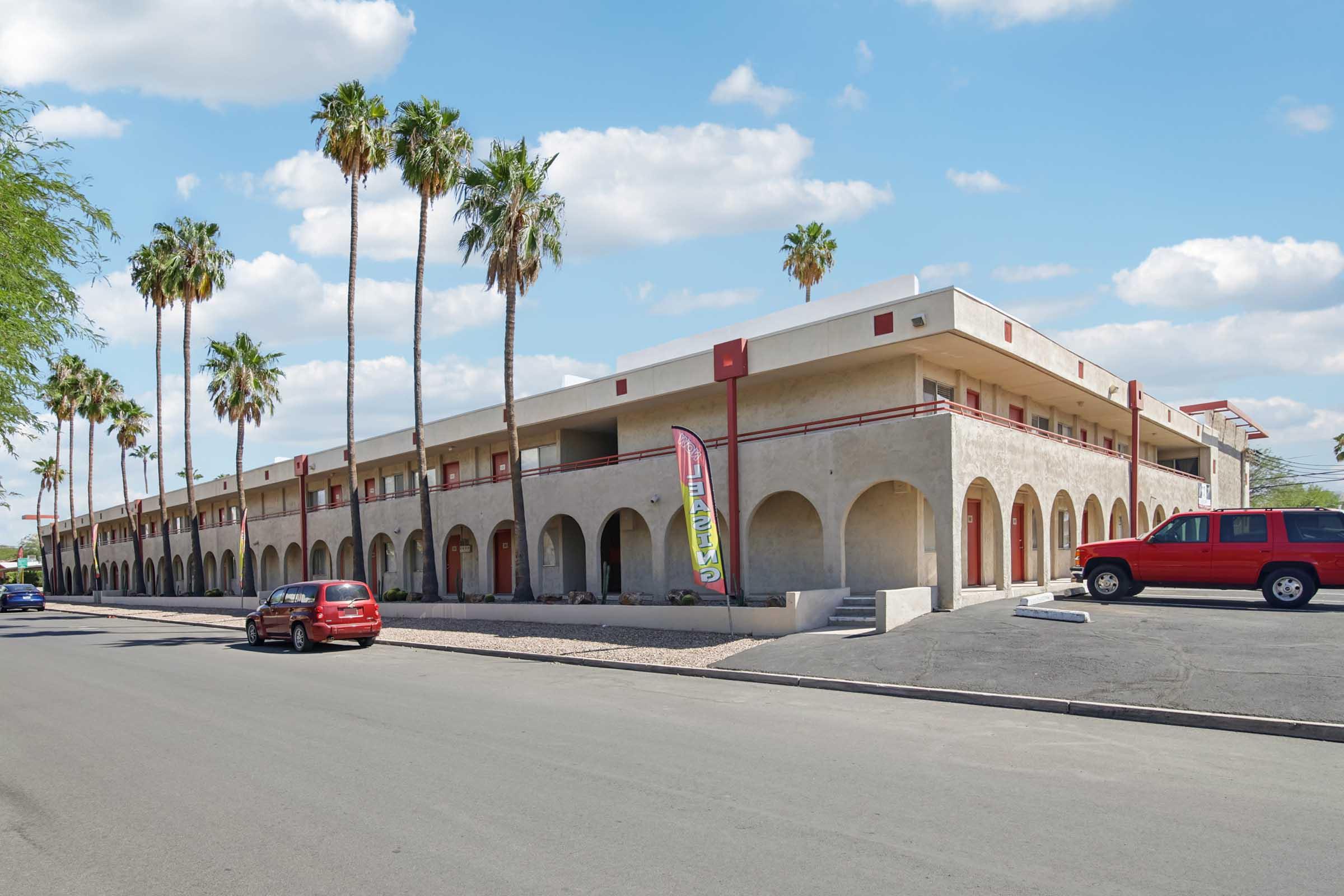 A low-rise building with an arched design, featuring multiple entrance doors, palm trees lining the front, and a bright blue sky with fluffy clouds. A red car is parked nearby, and a colorful banner is on display outside the building.