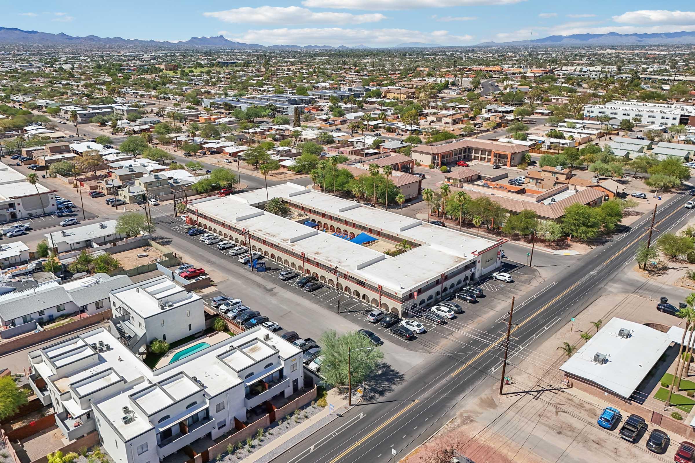 Aerial view of a suburban area featuring a shopping complex, surrounded by residential buildings and roads. The landscape includes a mix of single-family homes and apartments, with mountains visible in the background under a partly cloudy sky. Cars are parked in front of the shopping complex.