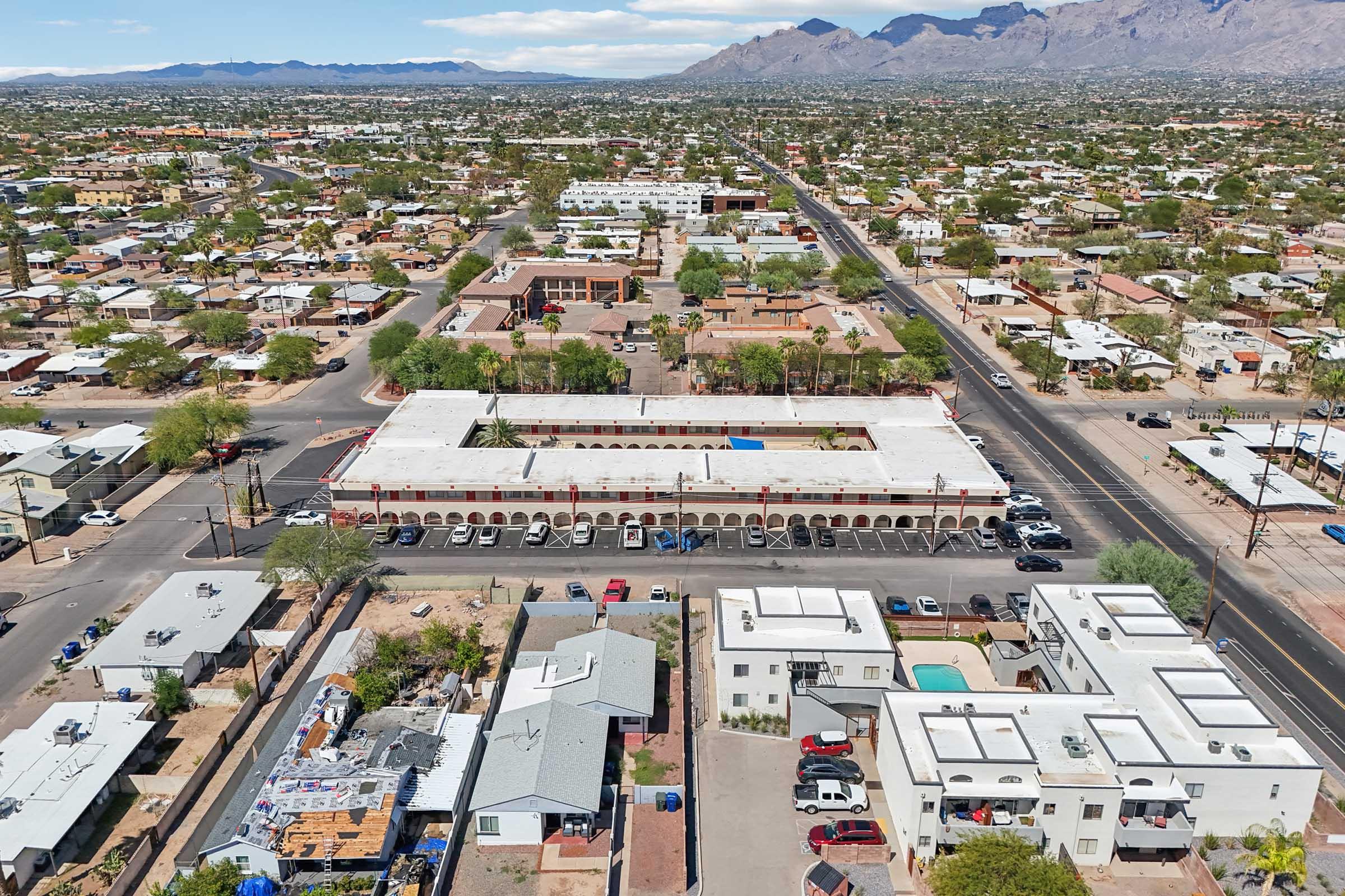 Aerial view of a suburban area featuring various residential buildings, streets, and green spaces. In the foreground, a low-rise structure with a flat roof is visible, surrounded by parking spaces. Mountains can be seen in the background, indicating a diverse landscape.