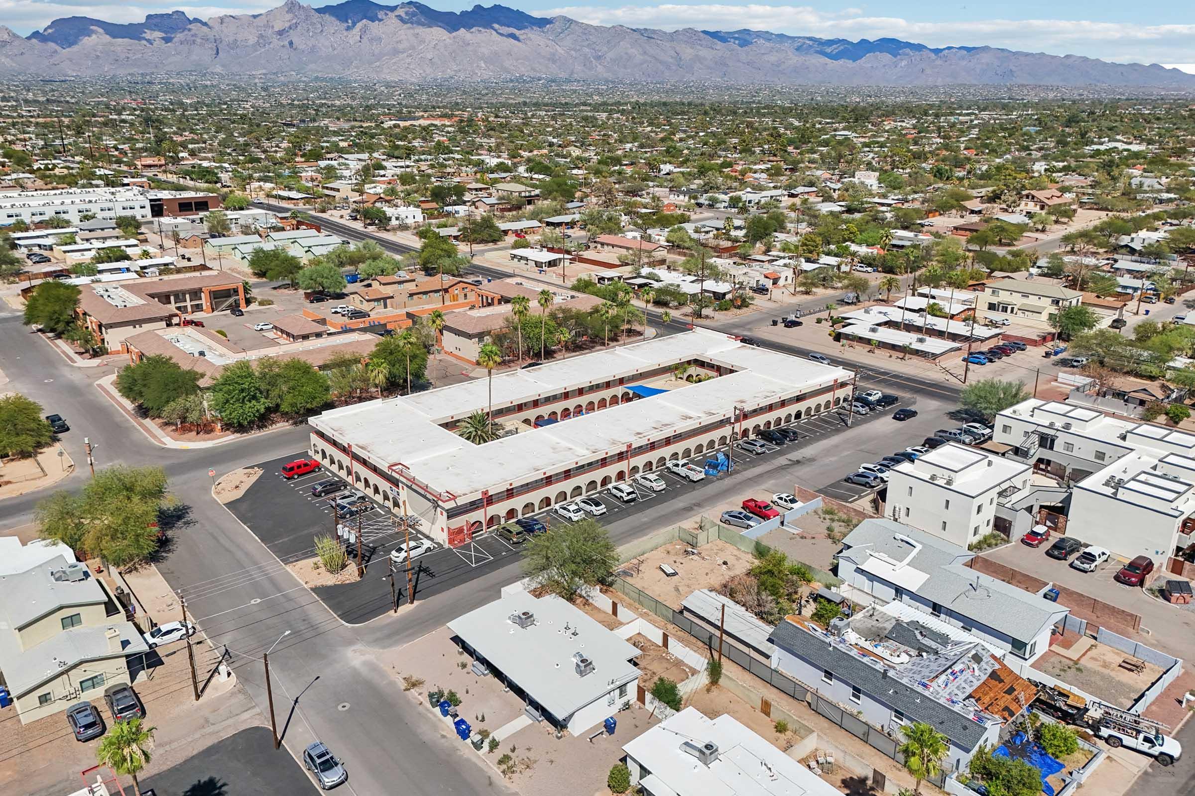 Aerial view of a suburban area featuring a large building with a red-tiled roof, surrounded by streets and smaller residential structures. The backdrop includes mountains under a blue sky with scattered clouds. Tree-lined streets and a mix of single-family homes and businesses are visible in the surrounding landscape.