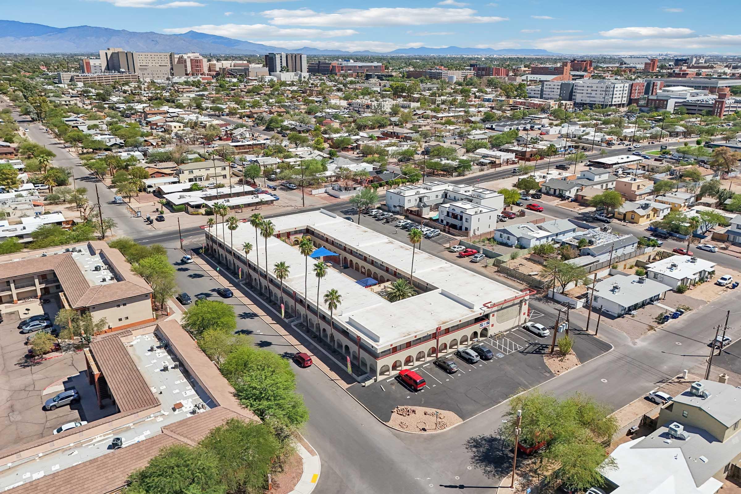 Aerial view of a downtown area featuring a mix of residential and commercial buildings, with palm trees lining streets. There are several low-rise structures, a parking lot with cars, and distant mountains under a clear blue sky. The scene captures the urban landscape and greenery.