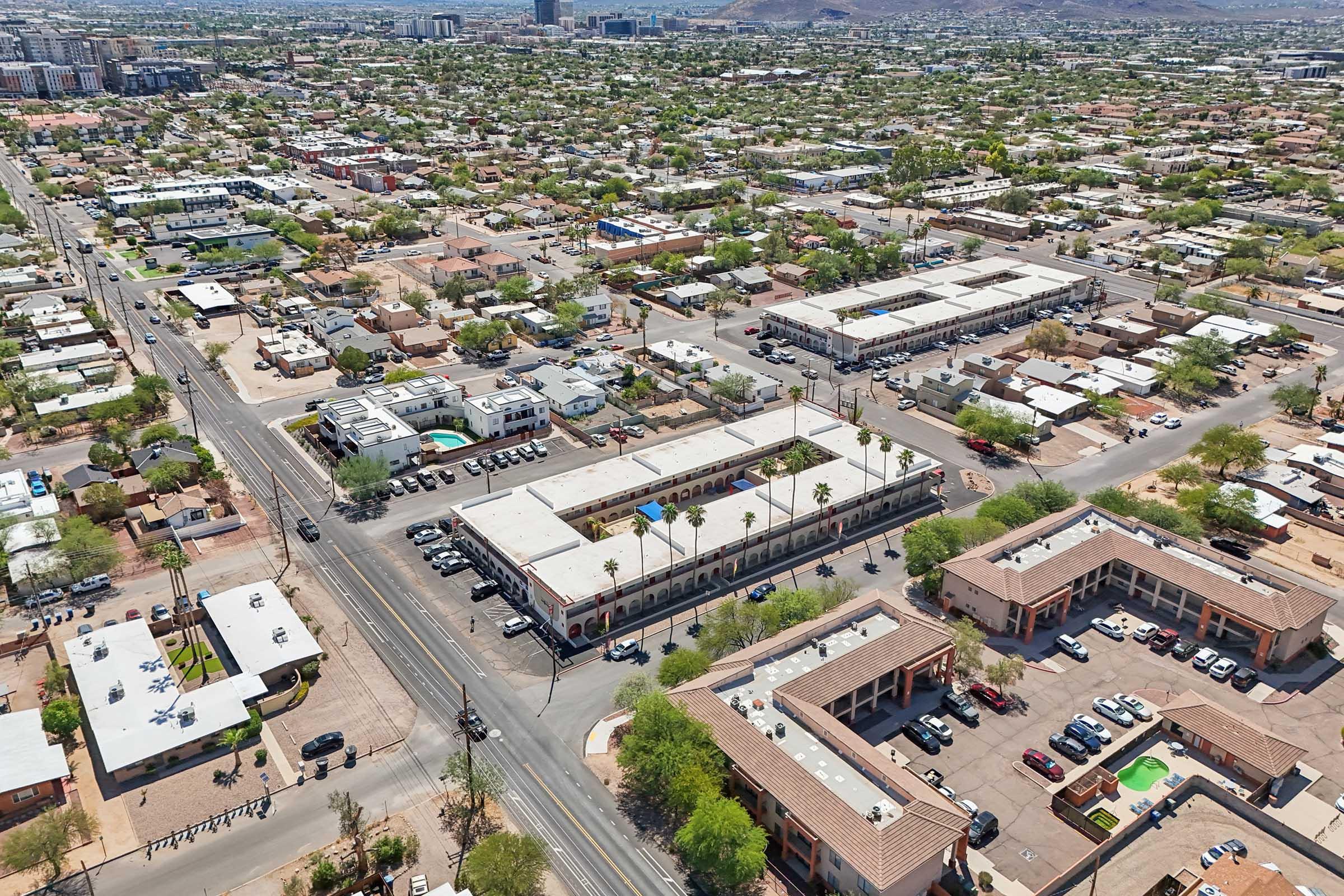 Aerial view of a suburban neighborhood featuring several low-rise buildings, including a motel, apartments, and single-family homes. The area is interconnected by roads, with greenery and palm trees visible. The background shows more urban development and hills in the distance.