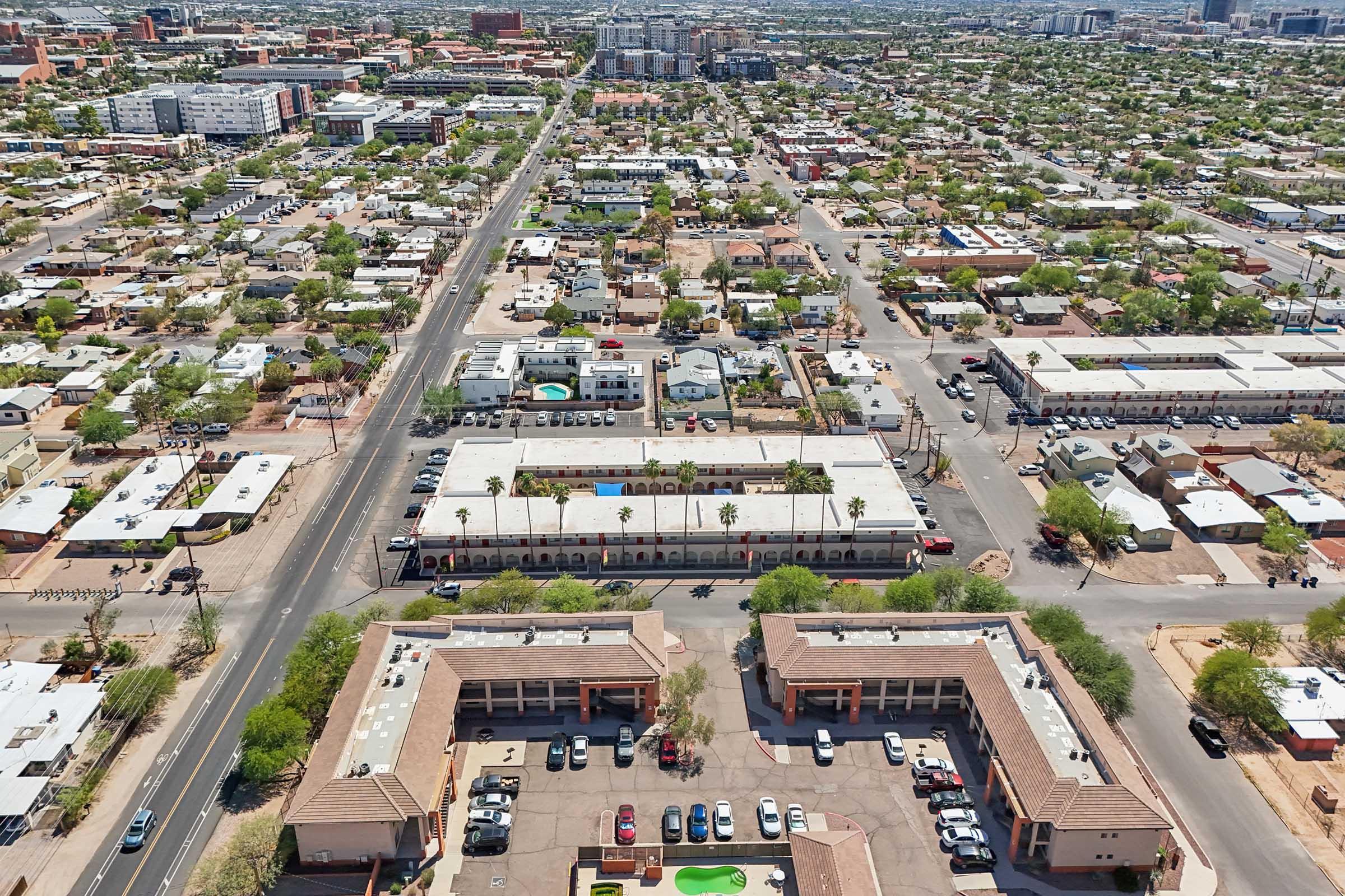 Aerial view of a suburban area featuring residential neighborhoods, commercial buildings, and a road. The landscape includes green trees and pools, with a mix of single-story and multi-story structures. The horizon shows city buildings in the distance beneath a clear sky.