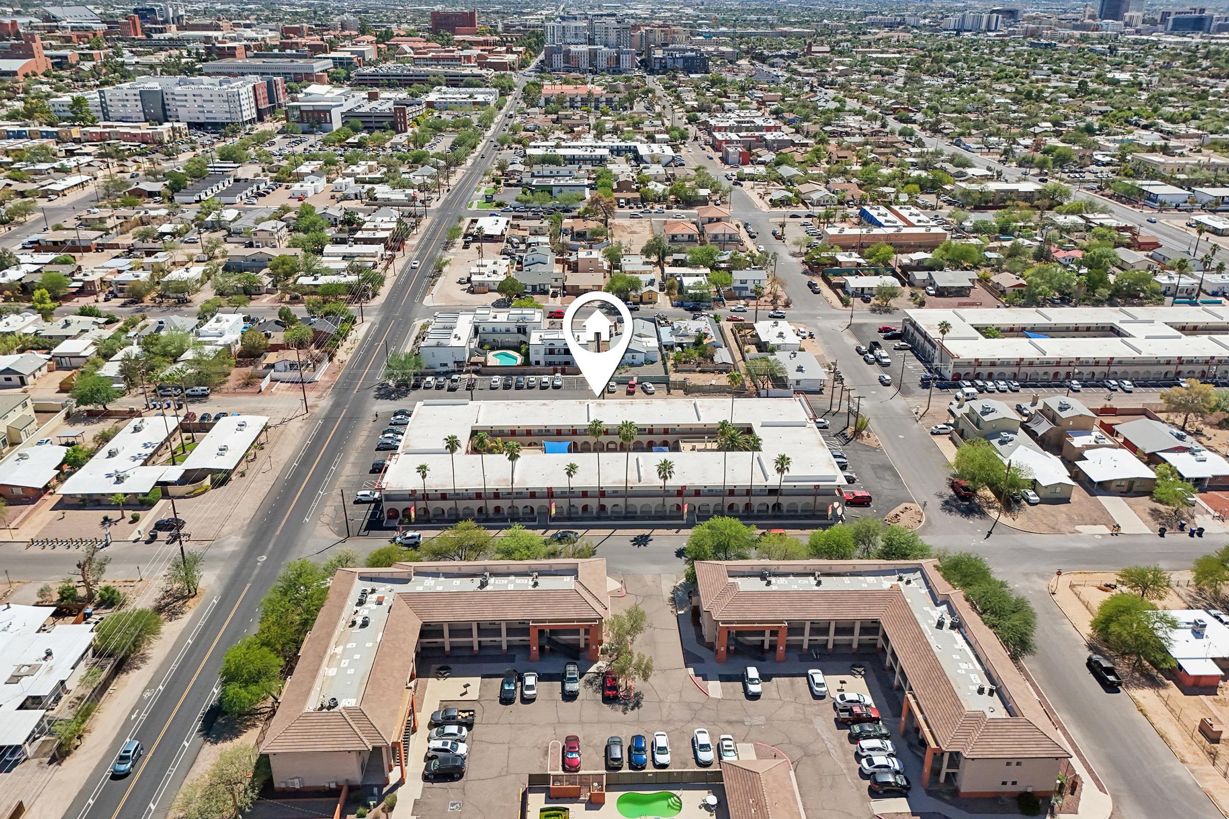 An aerial view of a neighborhood showing a residential area with several buildings, roads, and parking lots. A highlighted location marker indicates a specific building, which features a pool and palm trees. Surrounding structures consist of multiple homes and commercial units typical of urban settings.