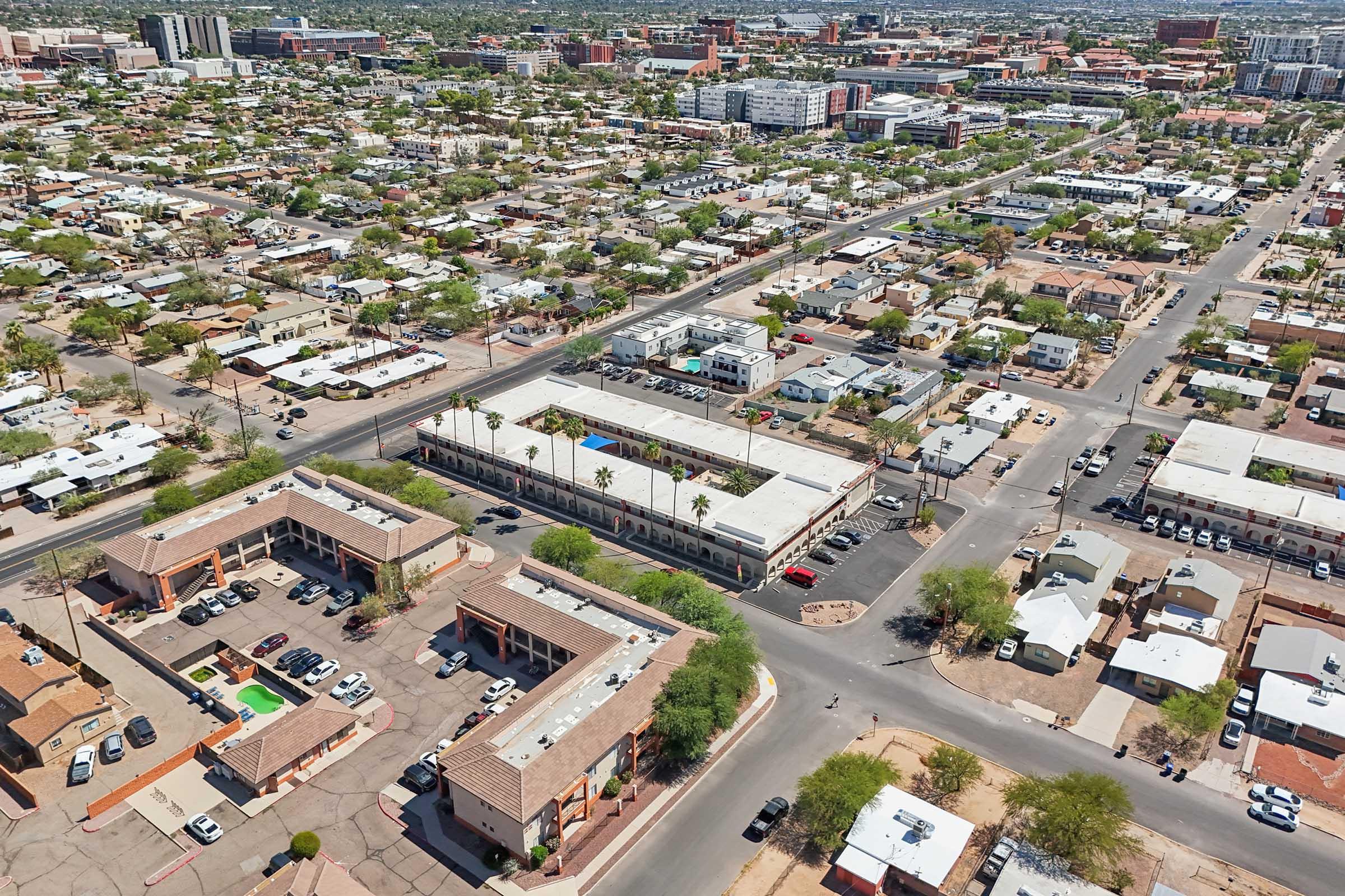 Aerial view of a suburban area featuring low-rise buildings, residential homes, and a mix of commercial structures. Several streets intersect, and parking lots are visible. The landscape includes patches of greenery amid predominantly urban development. In the background, taller buildings and a city skyline are visible.