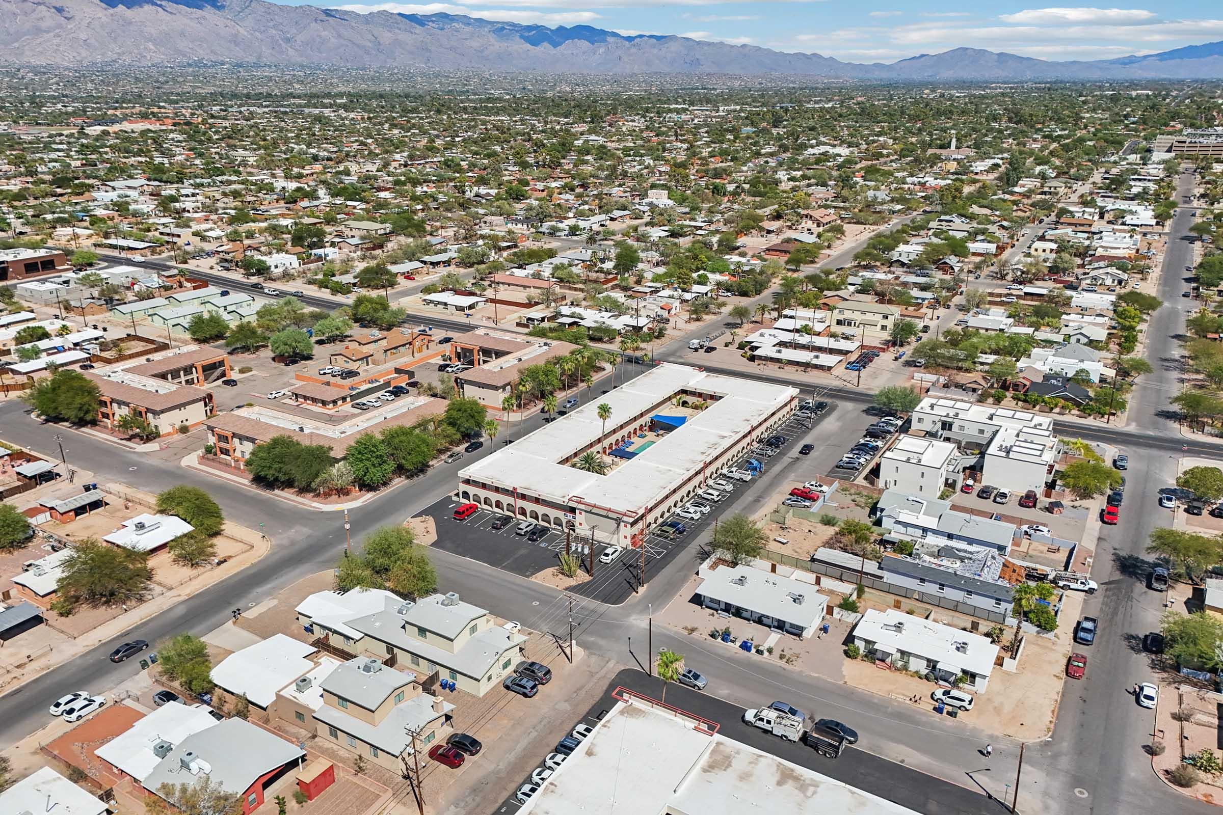 Aerial view of a suburban area featuring a grid of streets lined with single-story residential homes, commercial buildings, and a central complex with a pool. Surrounding mountains are visible in the background, and the landscape is lush with greenery, indicating a warm climate.