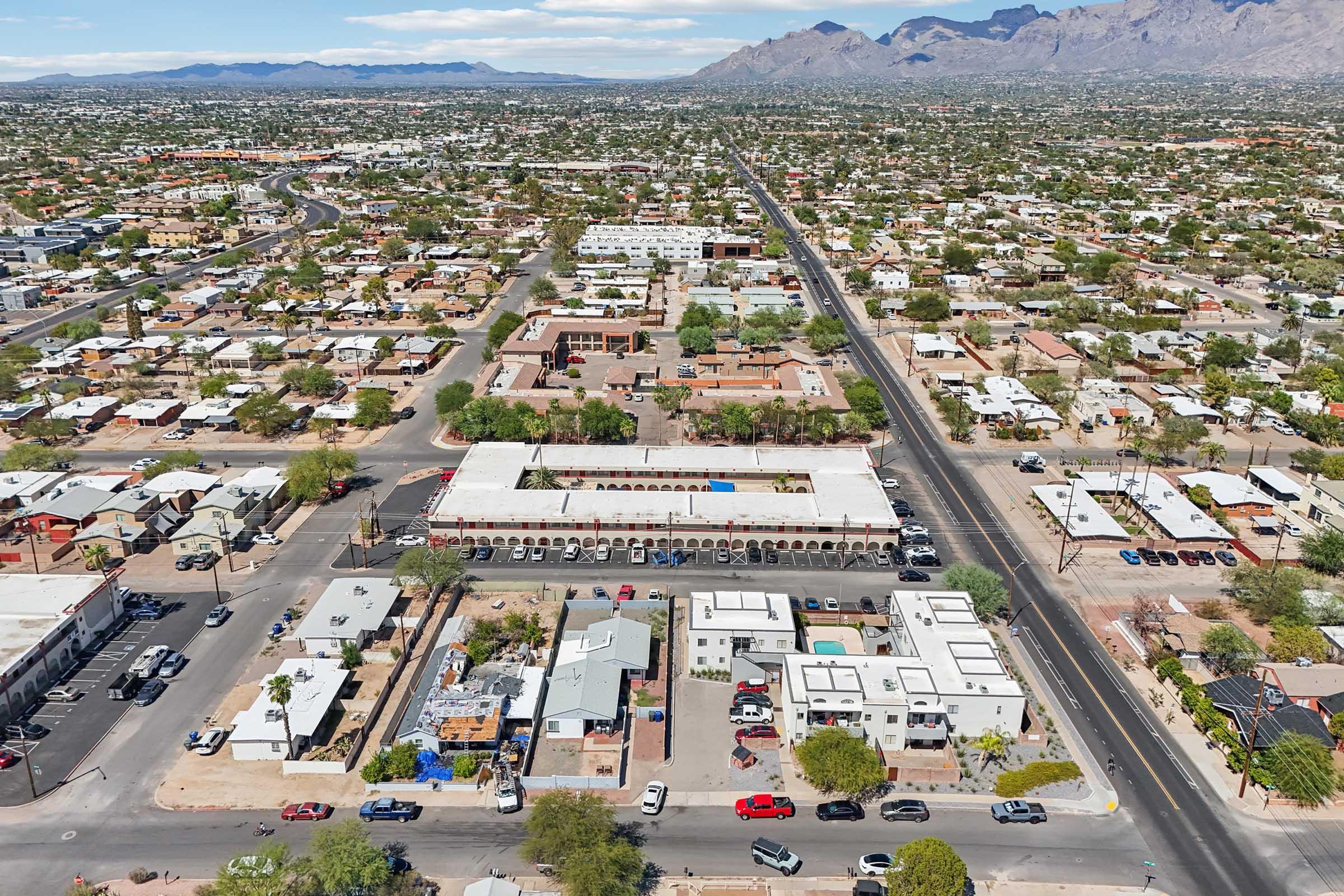 Aerial view of a suburban area featuring a grid layout of residential streets, shops, and businesses. Mountains are visible in the background, while various trees and vehicles are scattered throughout the neighborhoods. The scene captures a sunny day with clear skies.