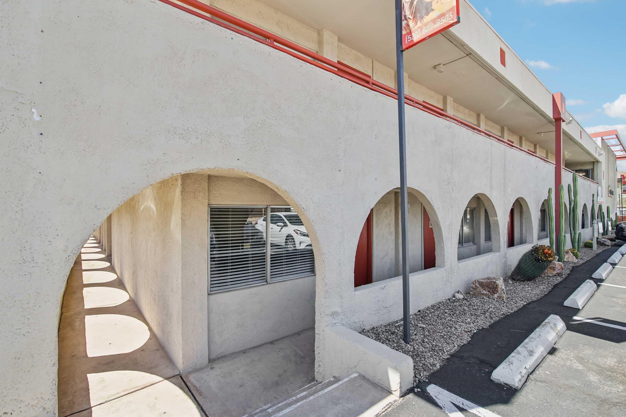 A view of a building's exterior featuring a series of arched openings along a tan wall. In the foreground, a parking area with white lines is partially visible, alongside decorative cacti and flower beds. The sky is bright with a few clouds, creating a sunny ambiance.