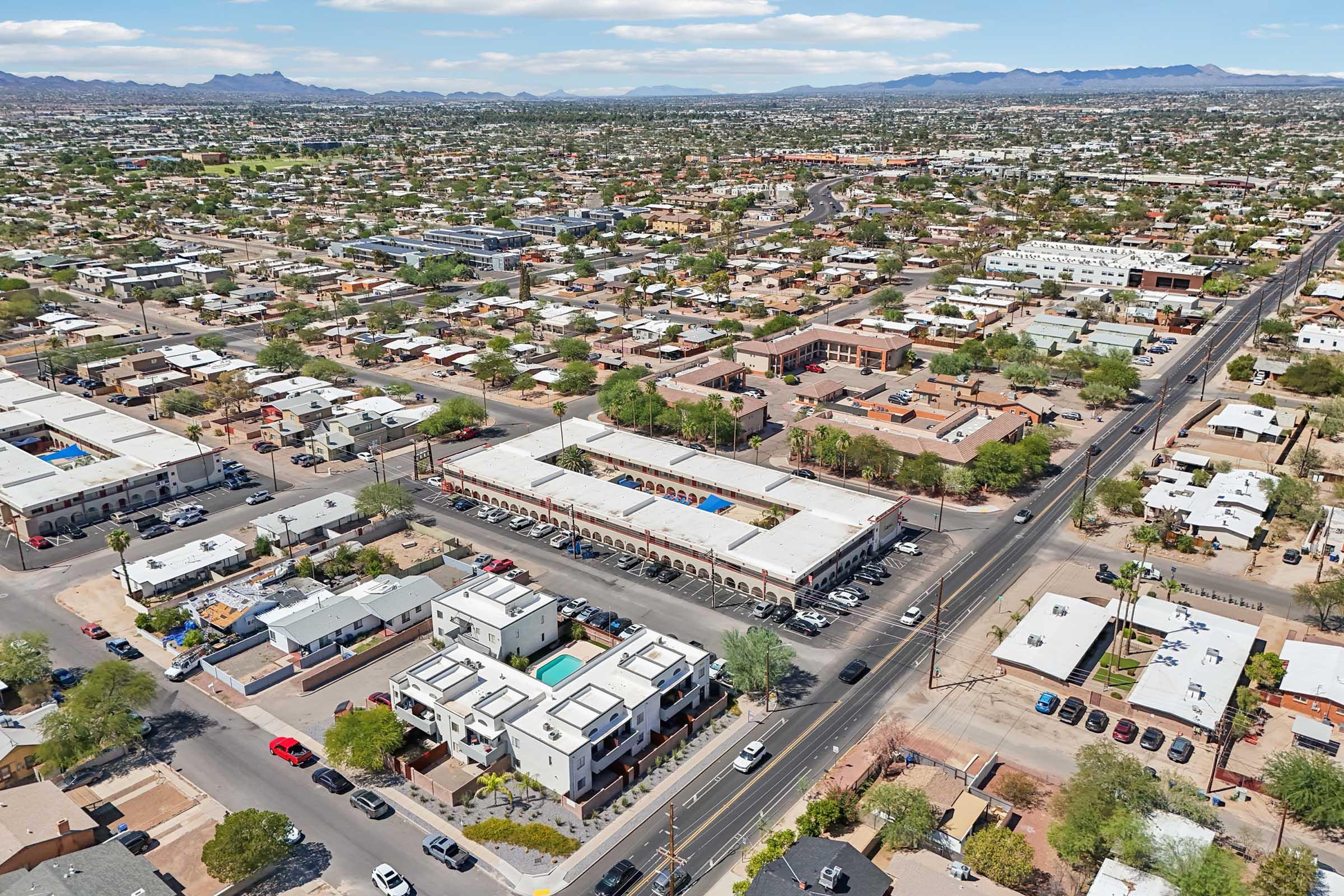 Aerial view of a densely populated urban area featuring a mix of residential and commercial buildings. Streets are lined with houses, and several swimming pools are visible in the complexes. Mountains are in the background under a partly cloudy sky.