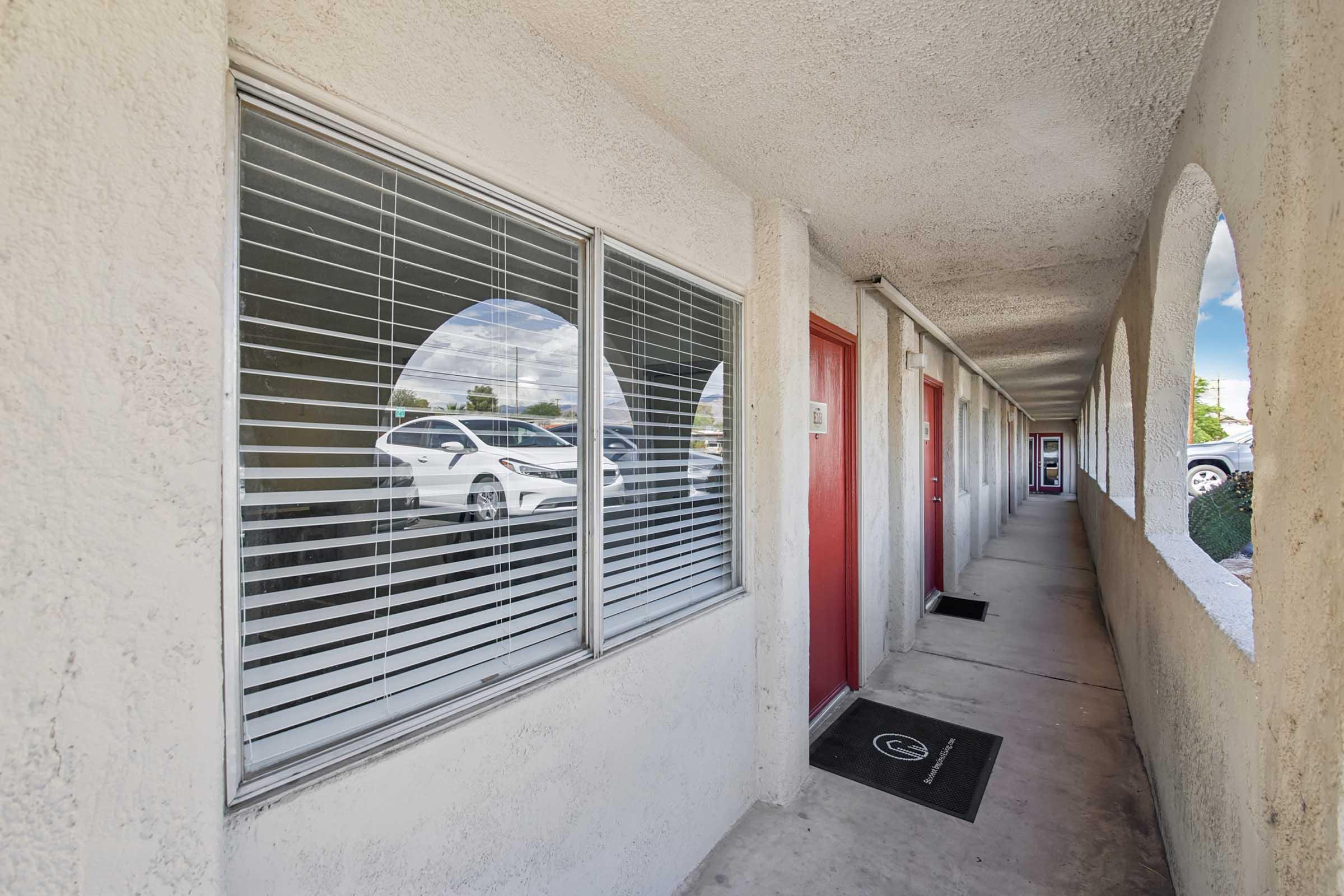 A corridor of a motel featuring several doors with red accents. Large windows with horizontal blinds are visible, allowing light to enter. The floor is concrete, and there is a welcome mat at the entrance of one of the rooms. The overall ambiance is casual and typical of budget accommodations.
