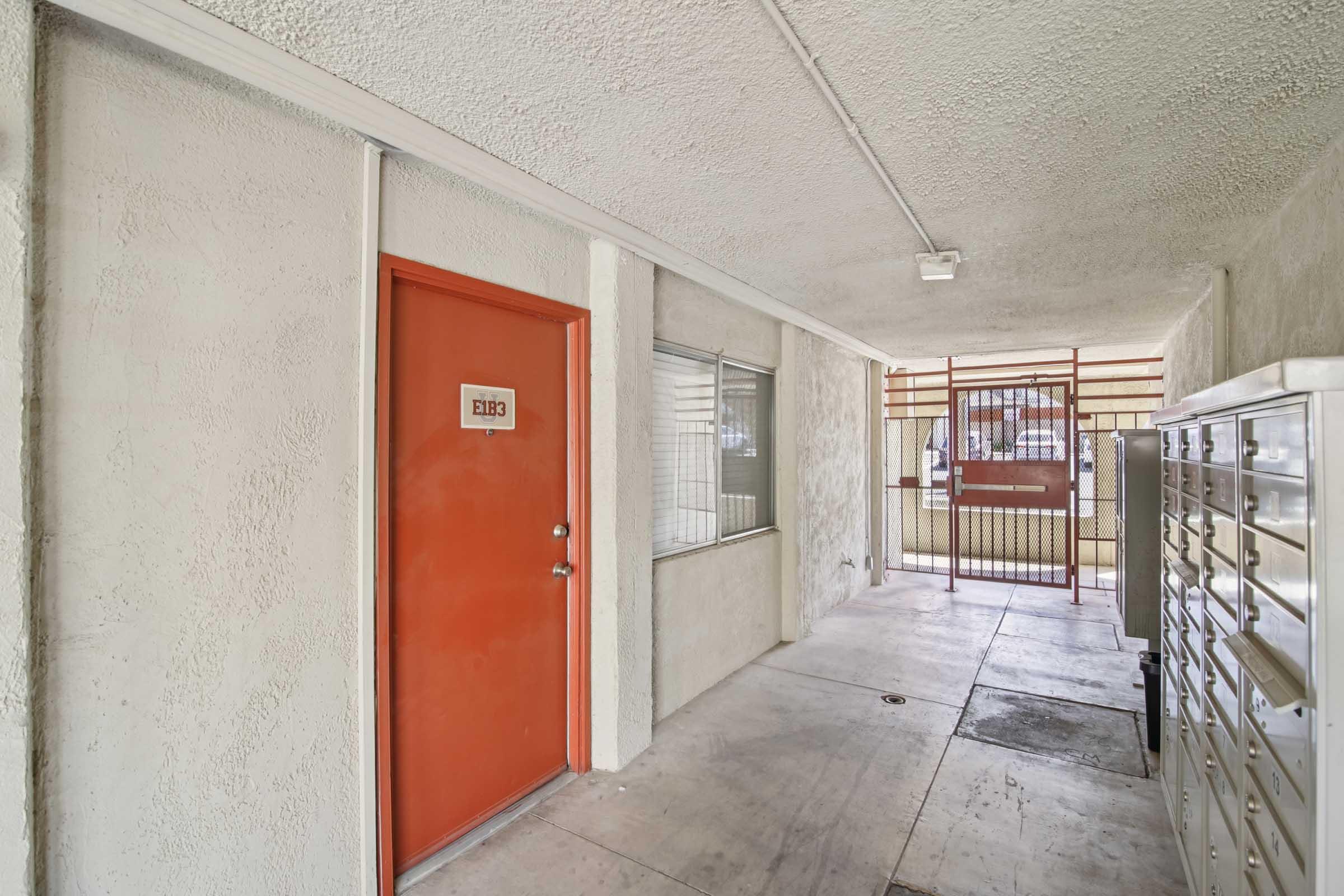 A view of a corridor in an apartment building. It features a door with the number "213," a window with blinds, and a set of mailboxes against the wall. The hallway has concrete flooring and a gated entrance visible in the background, providing a glimpse of the building's exterior.