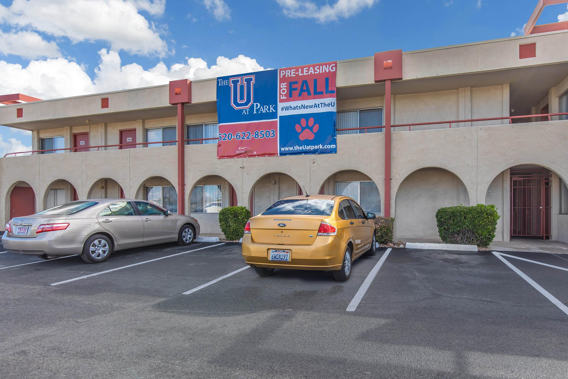 A parking lot view of a multi-story apartment building with a large banner advertising pre-leasing for fall. Two parked cars, a silver sedan and a yellow compact car, are visible in front of the building. The facade has a cream color with red accents and greenery along the base.