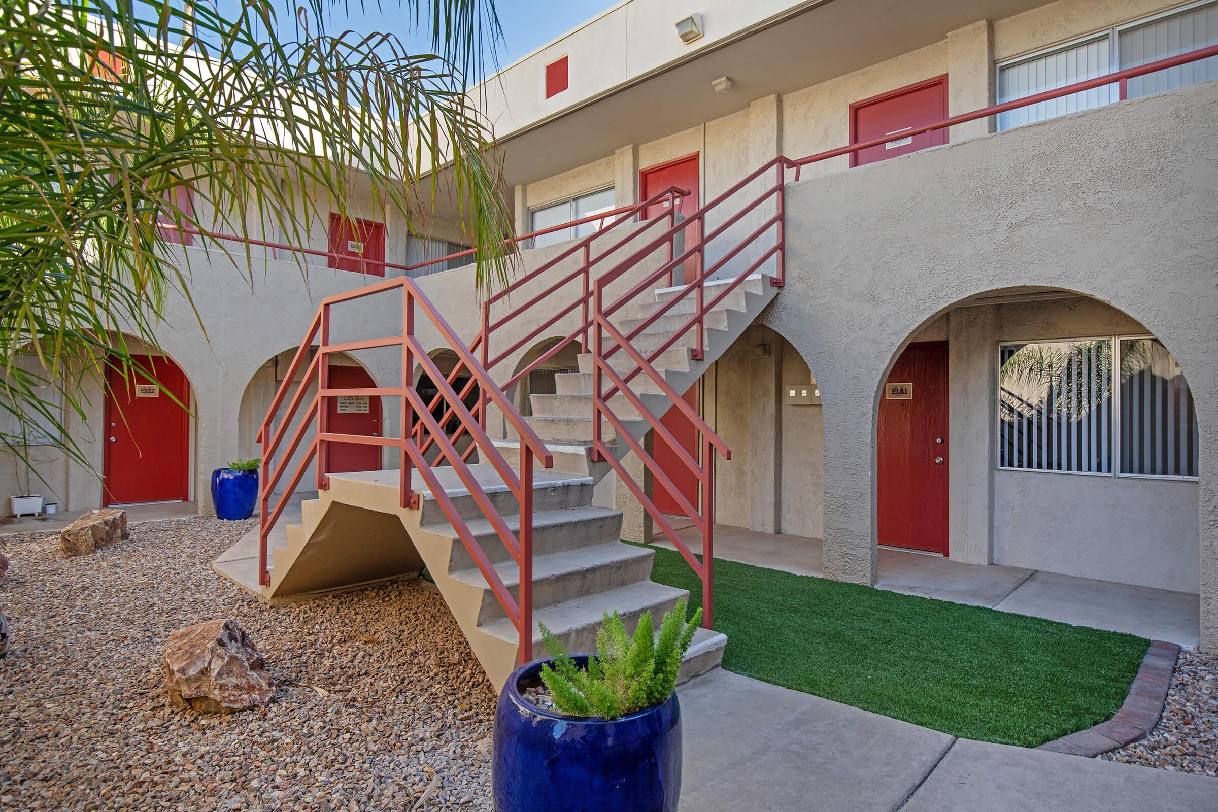 A view of a courtyard in an apartment complex featuring a staircase with red railings, surrounded by desert landscaping, including pebbles and a potted plant. The building has arched doorways and red doors, creating a vibrant and modern aesthetic.