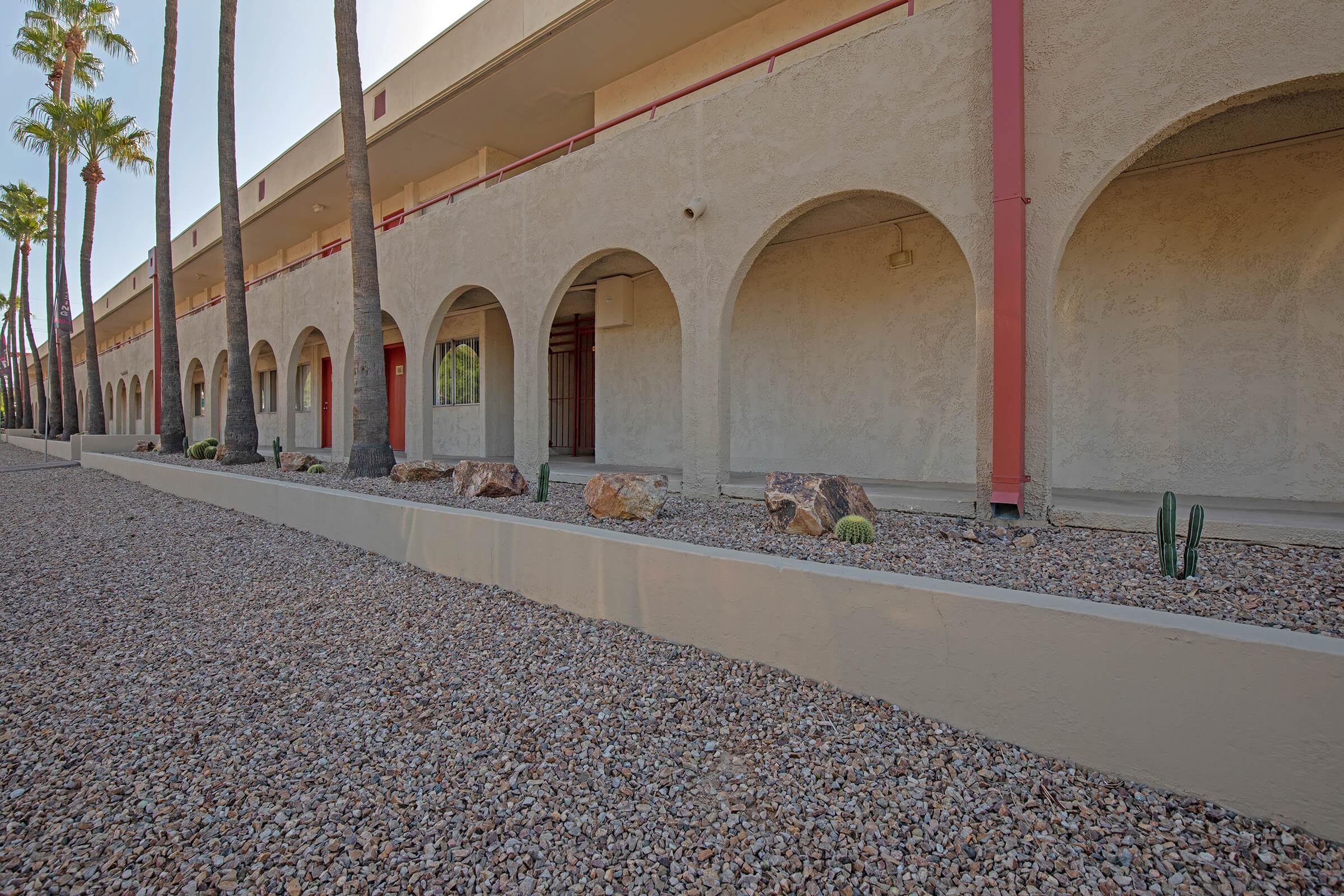 A view of a building facade featuring multiple arched entrances, surrounded by palm trees. The ground is covered with gravel, with a few large rocks and some small cacti visible along the base. The structure appears to be a low-rise complex, with a light-colored exterior and red accents.