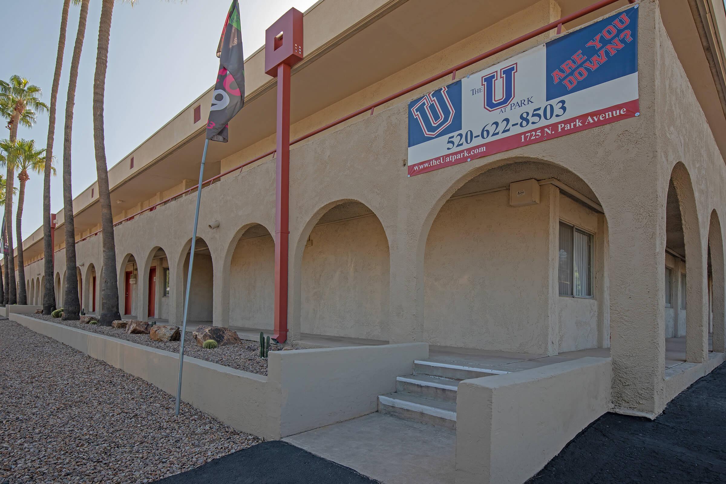 A view of a building with a stucco exterior featuring arches and a staircase. There are palm trees in the foreground and a sign that reads "U Park" along with a phone number. The overall setting appears to be in a warm climate, with gravel landscaping.