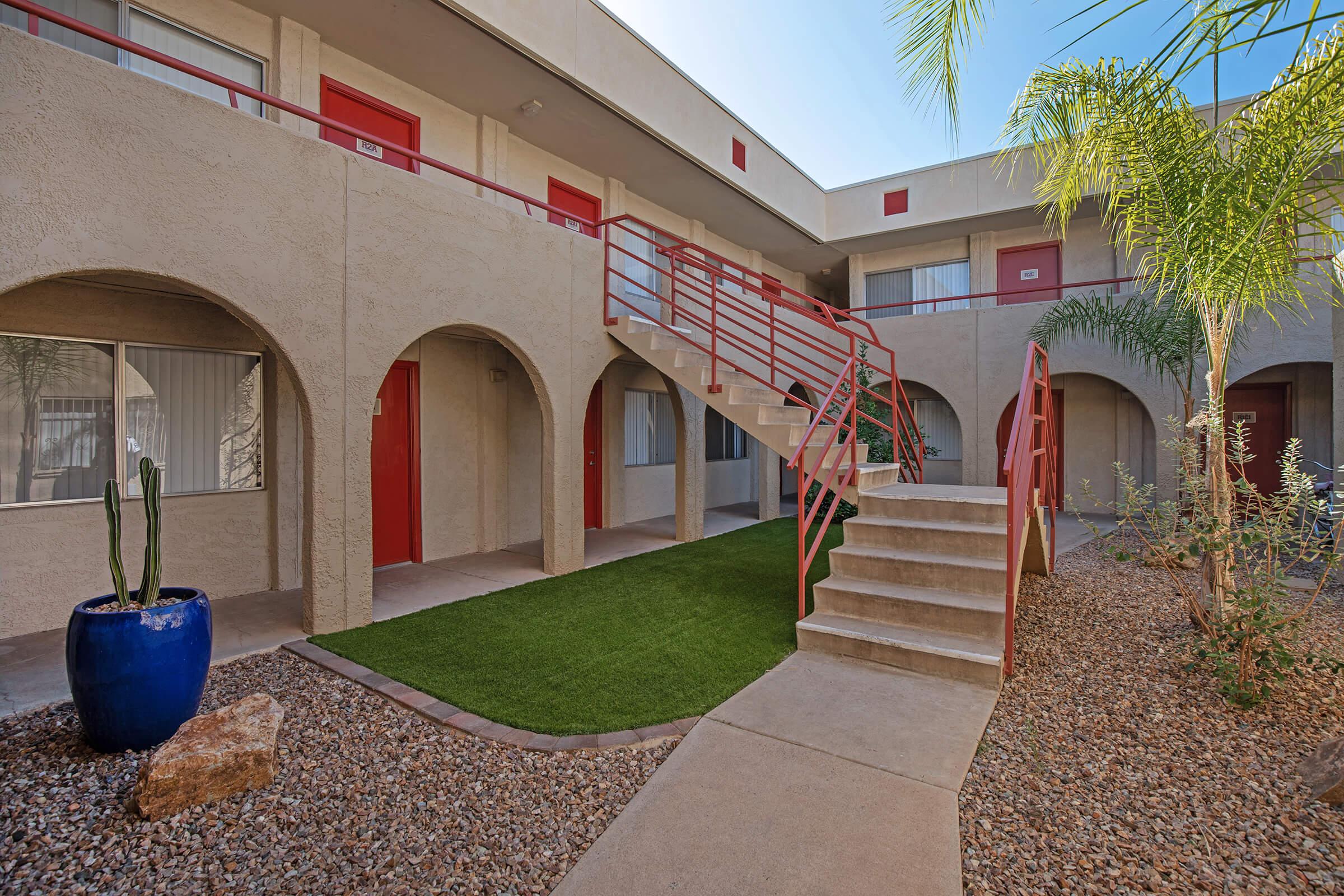 View of a courtyard in an apartment complex featuring arched doorways, bright red doors, a staircase with red railings, and artificial grass. Surrounding the area are potted plants, gravel landscaping, and palm trees under a clear blue sky.