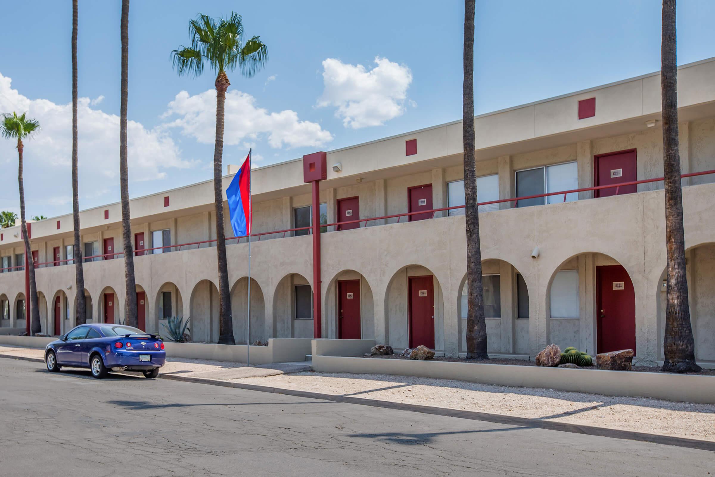 A row of two-story motel rooms featuring arched doorways, with red doors, palm trees lining the street, and a blue car parked nearby. A flag stands prominently in front of the building against a blue sky with some clouds. The setting appears to be a warm, sunny location.