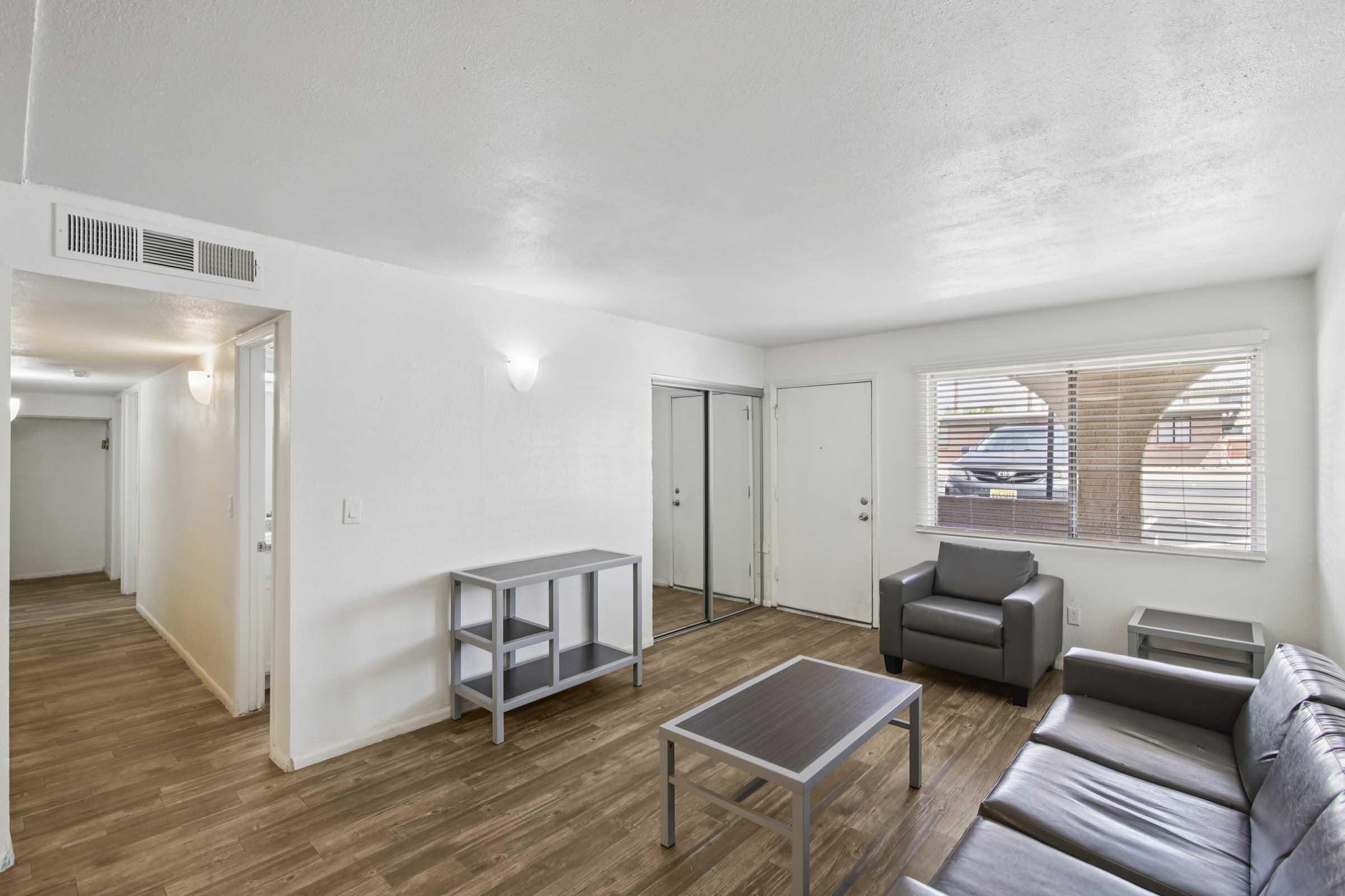 A bright, modern living room featuring a gray leather sofa, a glass-top coffee table, and a small metal shelf. Large windows provide natural light, and a door leads outside. The space has light-colored walls and a wood-like floor, creating a clean and welcoming atmosphere. A hallway is visible in the background.