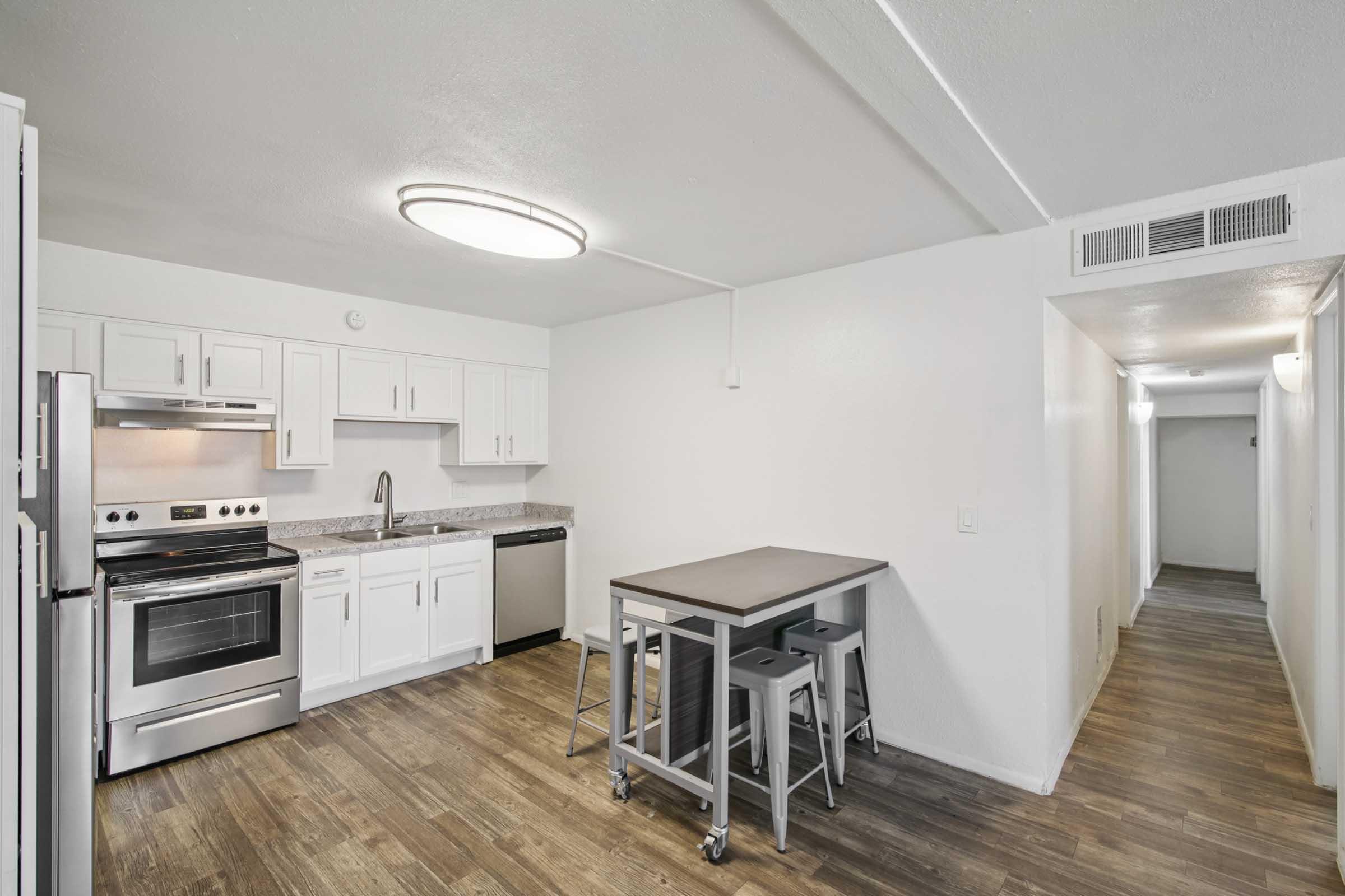Modern kitchen featuring white cabinets, stainless steel appliances including a refrigerator, stove, and dishwasher. A small dining table with metal stools is situated in the foreground, and the space has wooden flooring. A hallway is visible in the background, leading to another area.