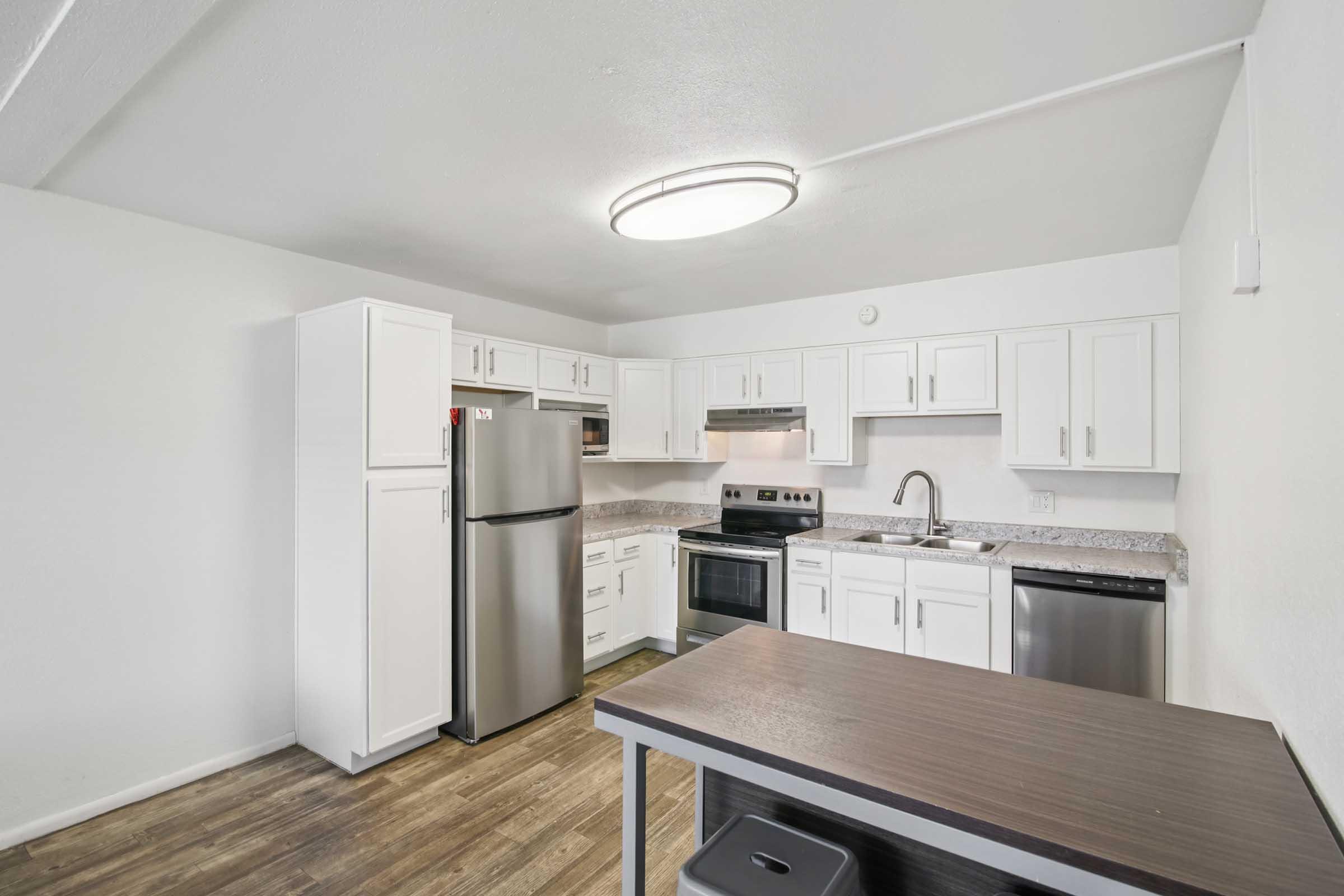 Modern kitchen featuring white cabinetry, stainless steel appliances including a refrigerator, stove, and dishwasher. A large, circular overhead light illuminates the space, which also has a wood laminate floor and a small table with a storage bin underneath. The walls are painted in a bright, neutral color.