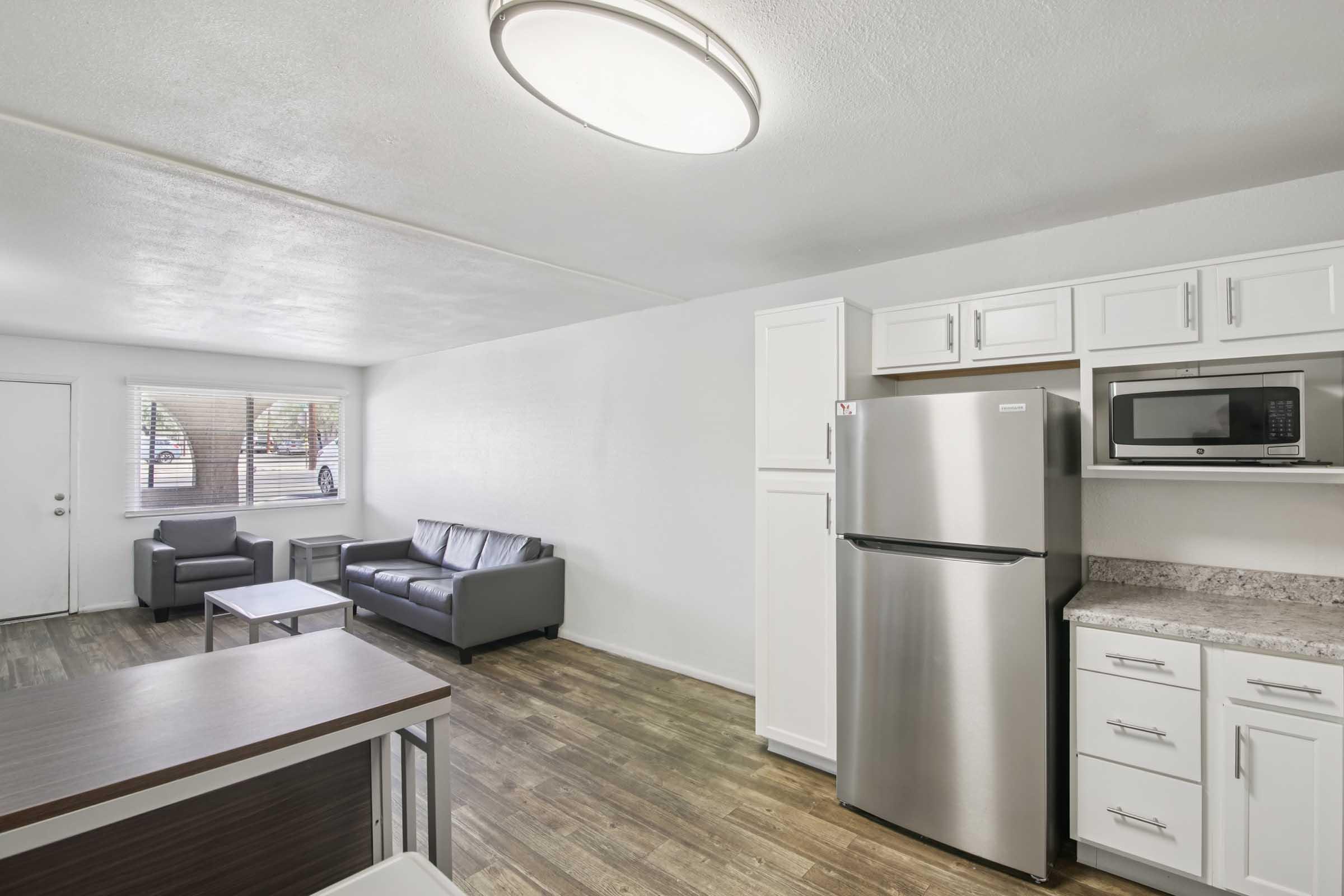 Interior view of a modern living space featuring a gray sofa, coffee table, and two chairs. The kitchen area includes a stainless steel refrigerator, microwave, and white cabinets. Natural light comes through a window, illuminating the room with a neutral color palette and wood-style flooring.
