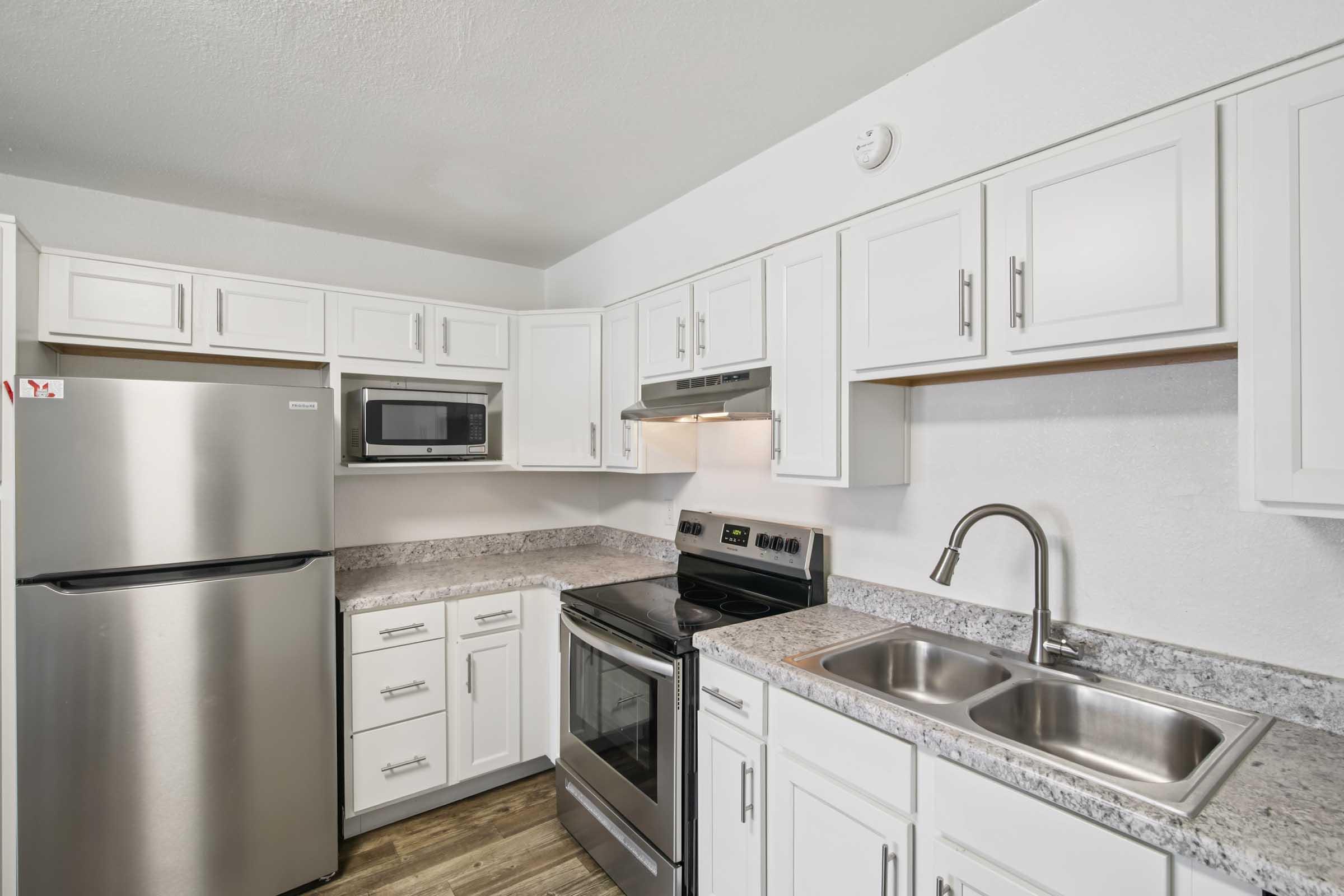Modern kitchen featuring white cabinetry, stainless steel appliances including a refrigerator, microwave, and stove, along with a double sink. The countertops are made of granite, and the flooring is wood-like laminate, creating a clean and functional design.