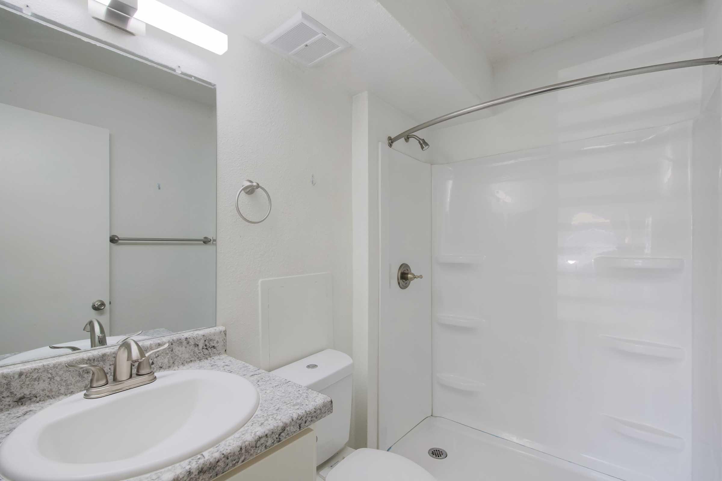 A clean and modern bathroom featuring a white shower stall, a small sink with a granite countertop, a toilet, and a mirror. The walls are painted white, and there is a towel rack and an overhead light. The space is well-lit and uncluttered, ideal for a minimalist aesthetic.