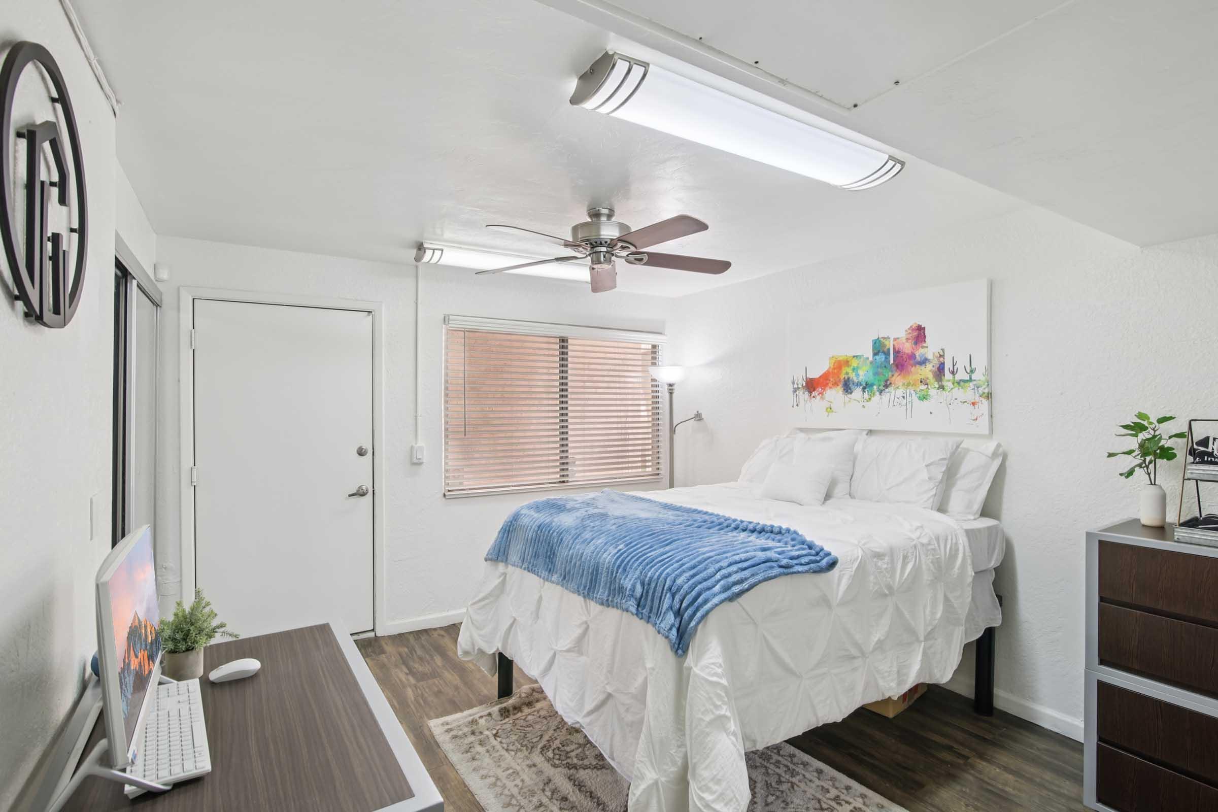 Brightly lit bedroom featuring a queen-sized bed with white linens and a blue throw blanket. There is a colorful abstract painting above the bed, a ceiling fan, a desk with a computer, and a window with blinds. The flooring is wood-style, and a small plant decorates the room.