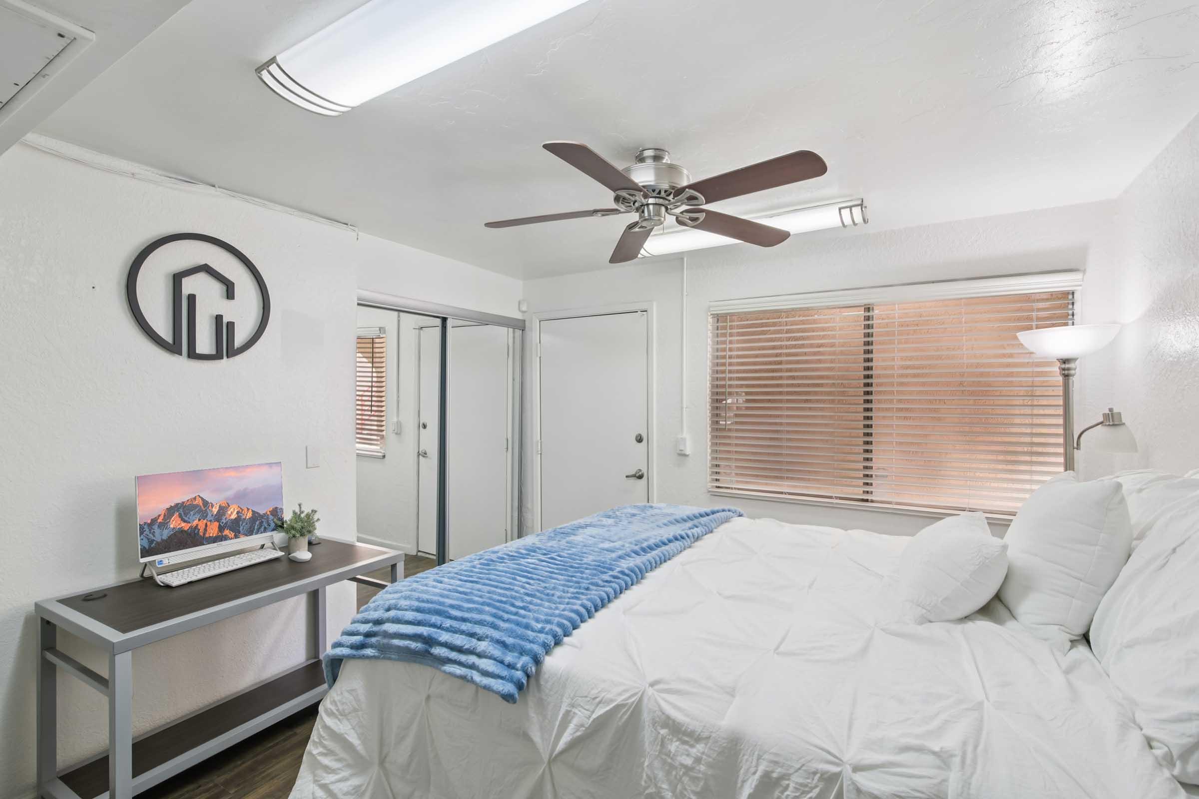 A cozy bedroom featuring a large bed with a white comforter and blue throw blanket. There's a ceiling fan, a small desk with a monitor displaying a mountain landscape, and a window with blinds. A minimalist décor includes a logo on the wall and natural light entering from the window.
