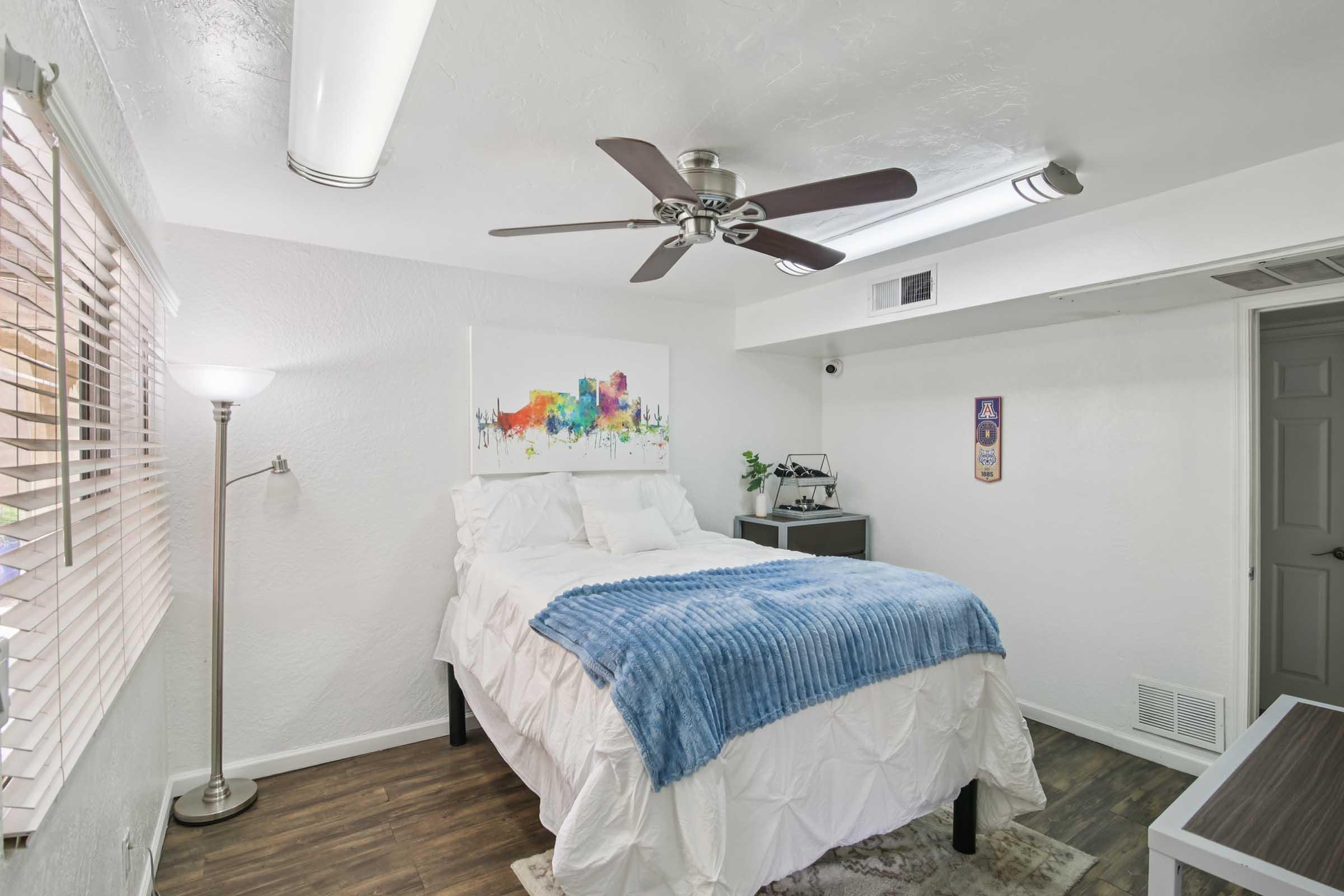 A well-lit bedroom featuring a queen-sized bed with a blue throw blanket, a modern ceiling fan, and wooden flooring. There is a decorative wall art piece with colorful cityscape, a floor lamp, and a small desk area visible. Sunlight streams through partially closed blinds.
