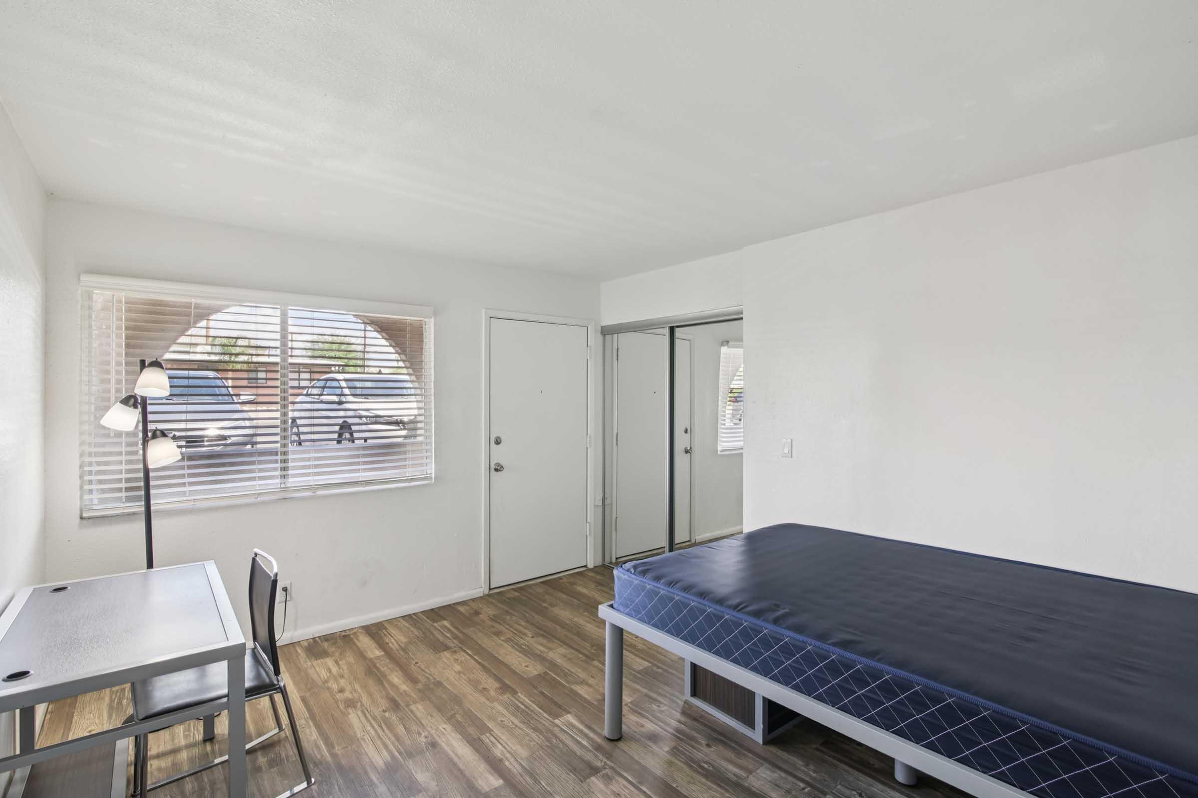 A sparsely furnished room featuring a single bed with a dark blue mattress, a desk with a chair, and a window with blinds. The walls are painted white, and there is a door leading outside. The flooring is wooden.