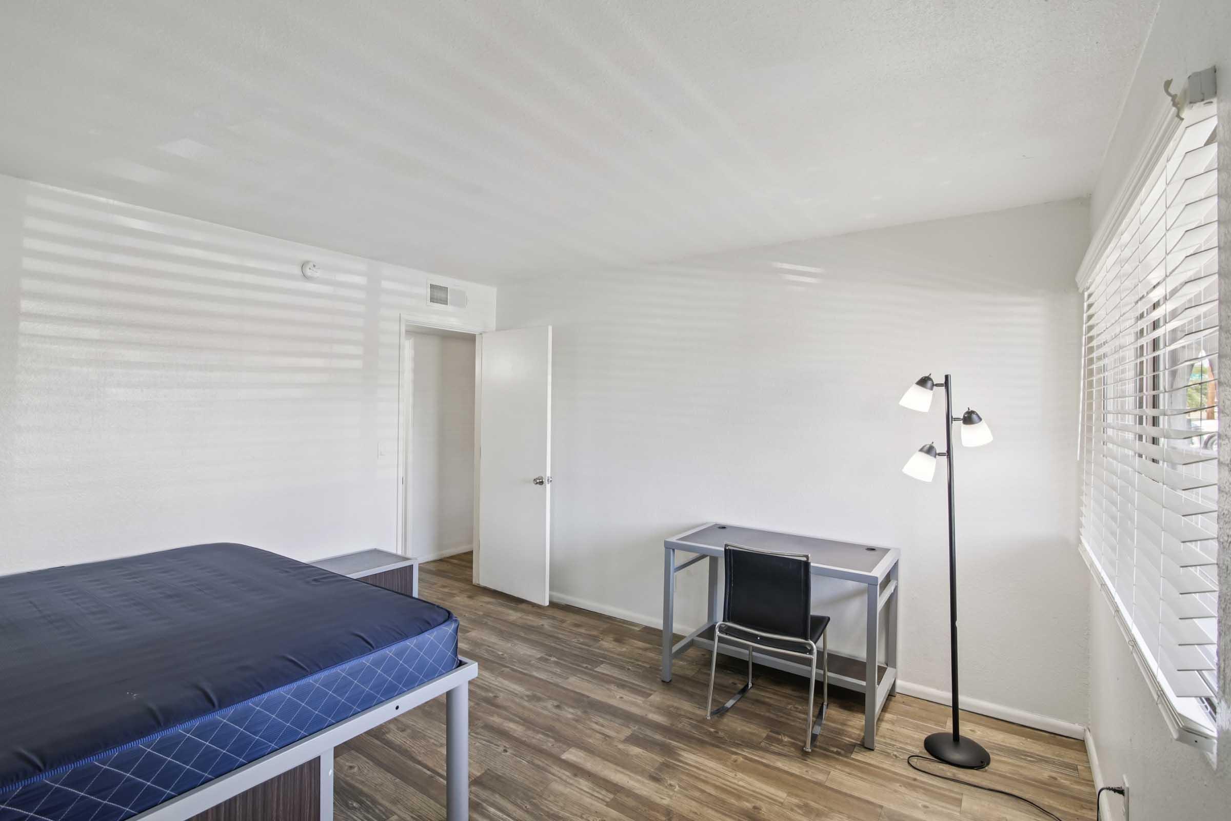 A minimalistic bedroom featuring a bed with blue bedding, a desk with a black chair, and a floor lamp. The room has light-colored walls and wooden flooring, with natural light streaming through white blinds. A closed door is visible in the background.
