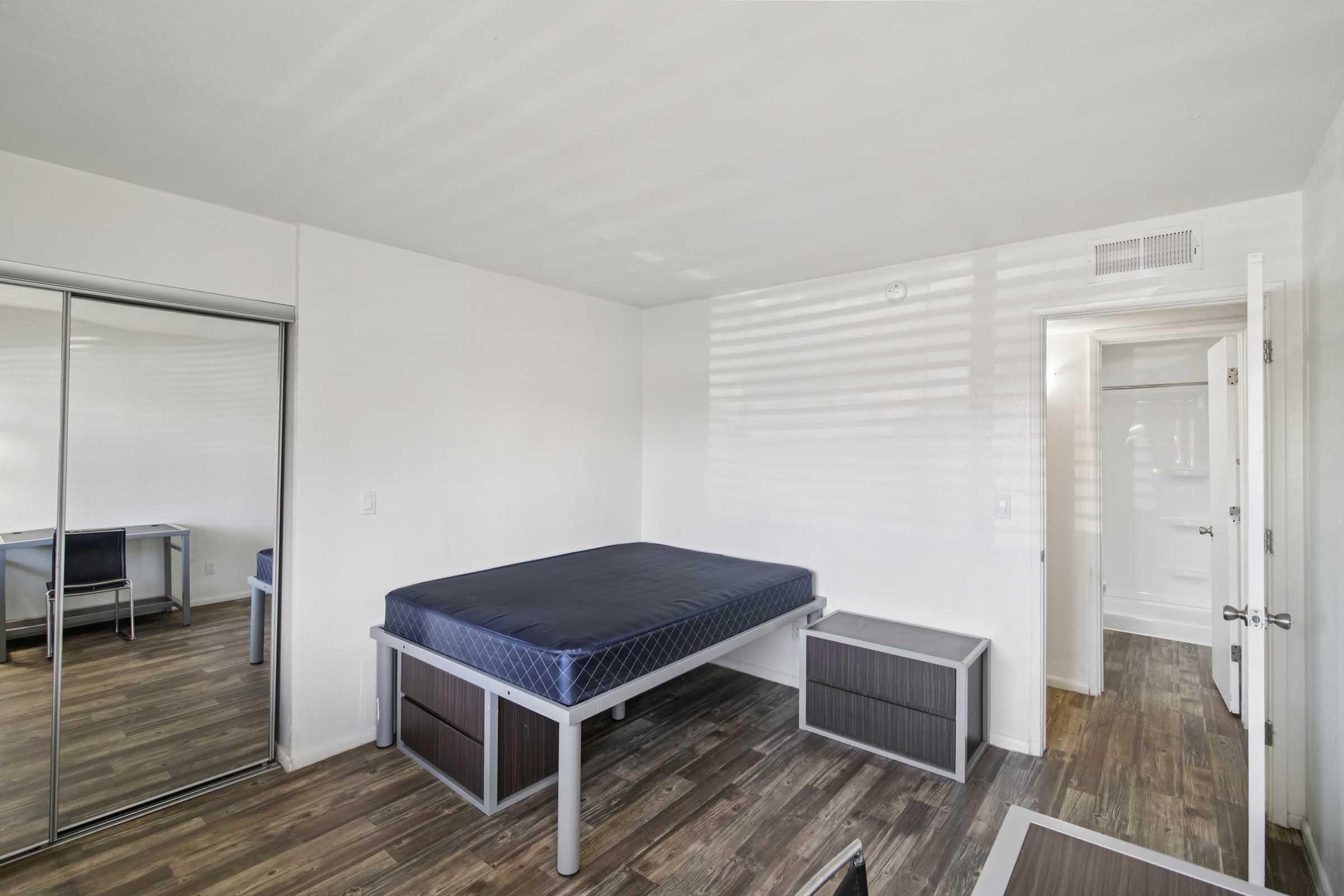 A minimalist bedroom featuring a bed with a blue mattress, a nightstand, and a large mirror closet. The walls are white, and the floor has wood-like laminate. Natural light casts soft shadows, enhancing the simplicity of the space. A door leads out of the room, visible in the background.