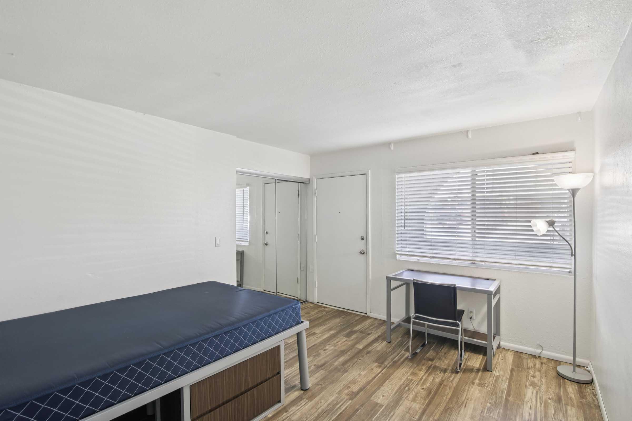 A simple, minimalist room featuring a single bed with a blue mattress, a desk and chair, and a floor lamp. A large window with blinds allows natural light, and a closet space is visible. The flooring is light wood, creating a clean and tidy atmosphere.