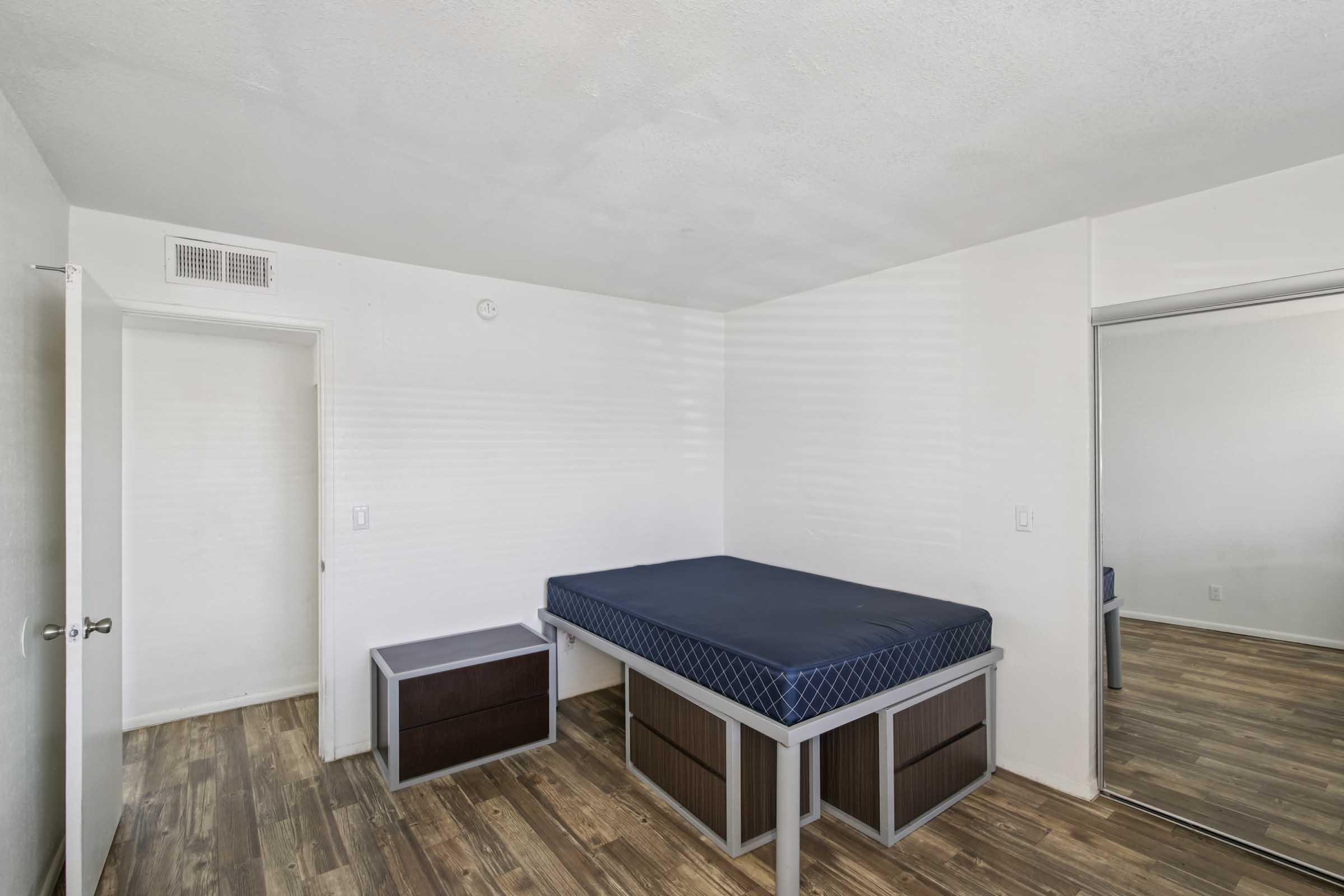 A simple, unadorned bedroom featuring light-colored walls and wooden flooring. It contains a bed with a mattress and a bedside table. A closet with mirrored doors is visible on the right side, and a closed door is seen in the background. The room is well-lit and appears tidy.