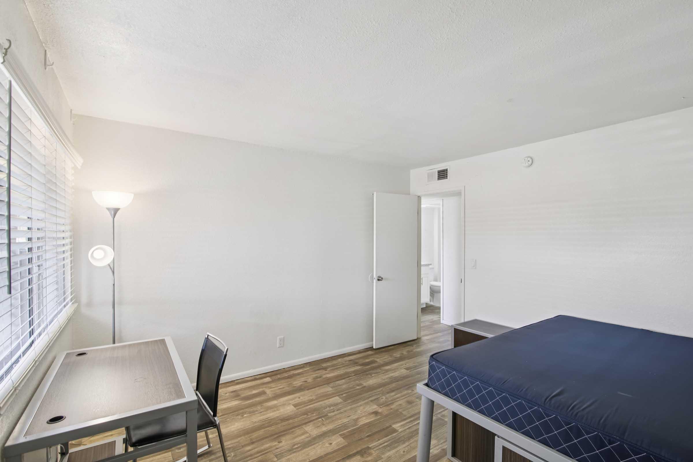 A simple, minimalist bedroom featuring a bed with a blue mattress, a desk with a chair, and a floor lamp. The walls are painted white, and there are light wood laminate floors. A window with blinds allows natural light, and a partially open door leads to another room.