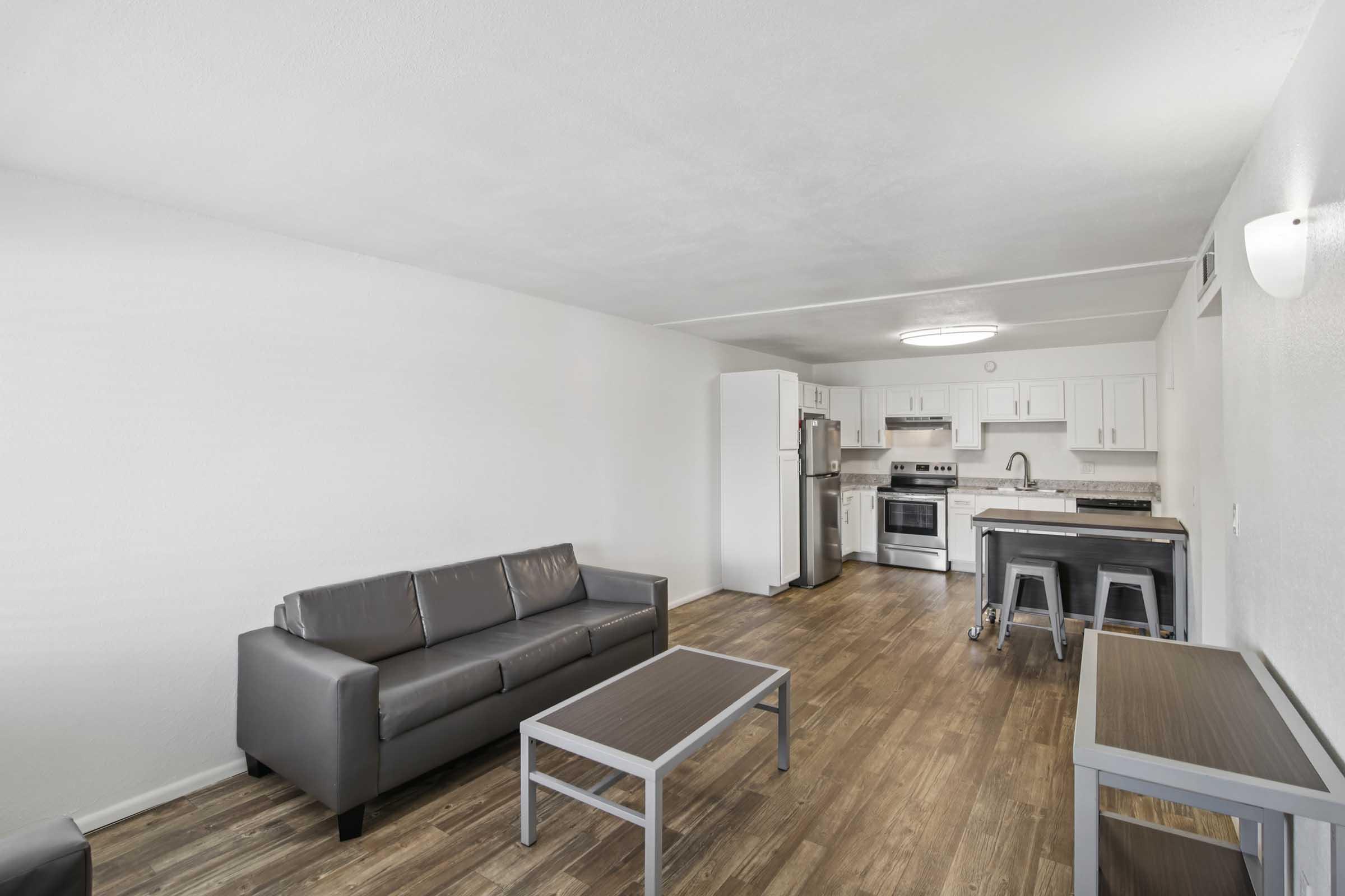 Modern living space featuring a gray couch and a wooden coffee table. The kitchen area includes white cabinets, stainless steel appliances, and a countertop with bar stools. The room has neutral-colored walls and wooden flooring, creating a spacious and bright atmosphere.