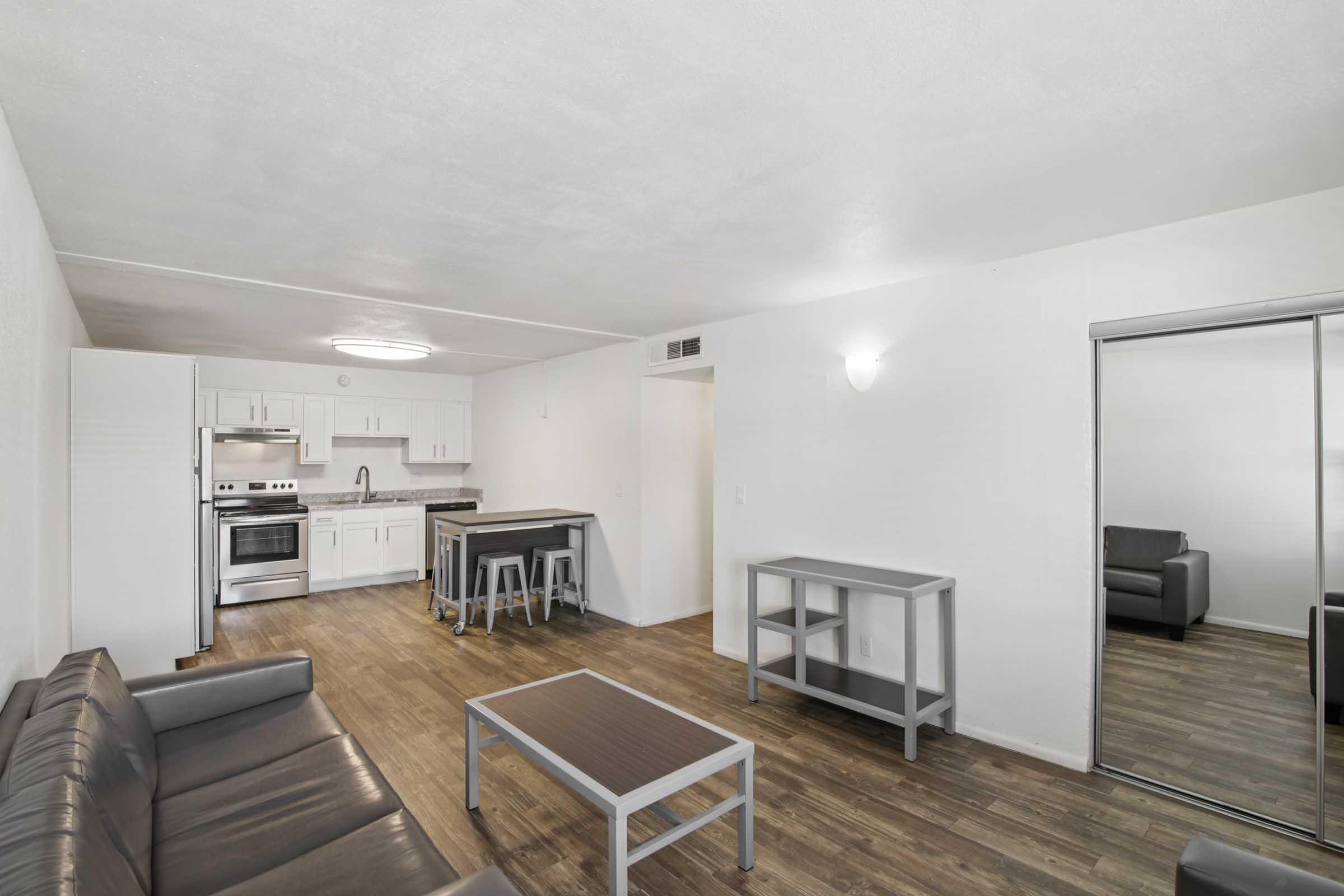 Interior of a modern living space featuring a kitchen area with stainless steel appliances, a dining table with bar stools, and a seating area with a dark couch and coffee table. The space has light-colored walls and wood-like flooring, creating a bright and open atmosphere. A mirrored closet door is visible in the background.