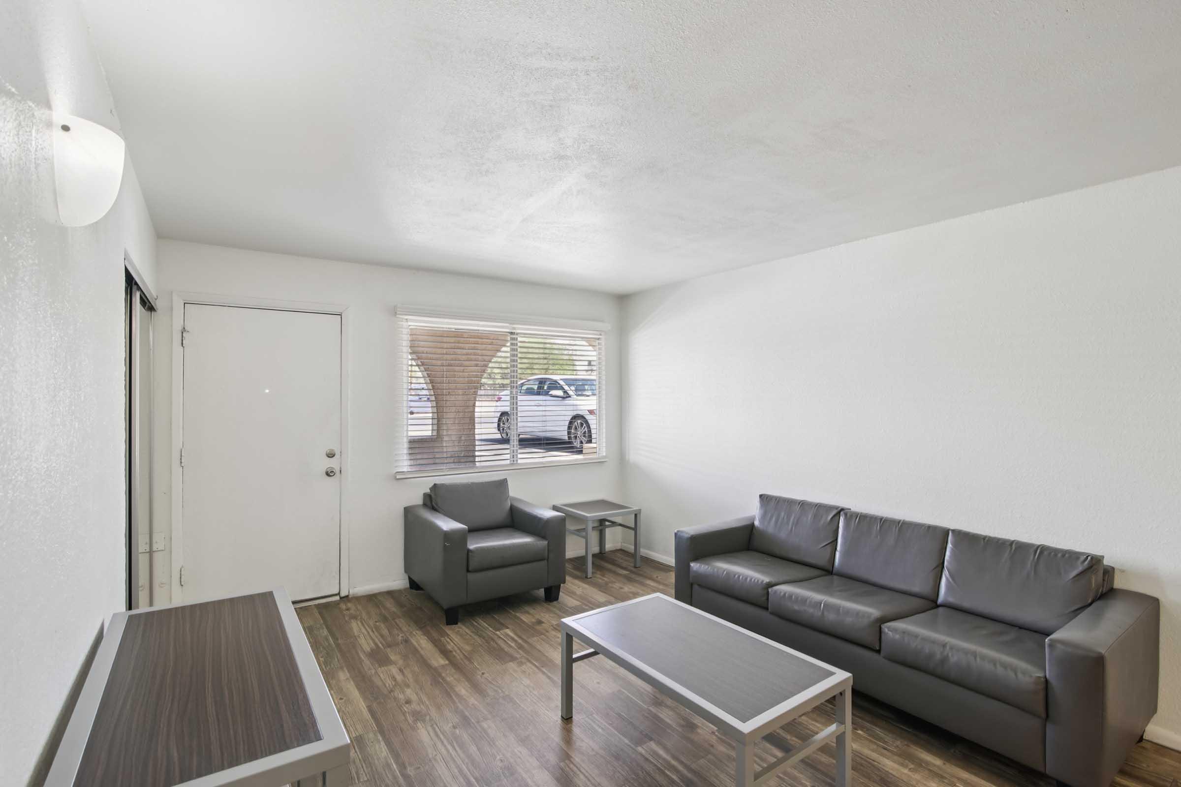 A simple, modern living room featuring a gray sofa, two gray armchairs, and a rectangular coffee table on wooden flooring. Natural light streams in through a window with blinds, revealing a glimpse of a car parked outside. The walls are painted white, creating a bright, open atmosphere.