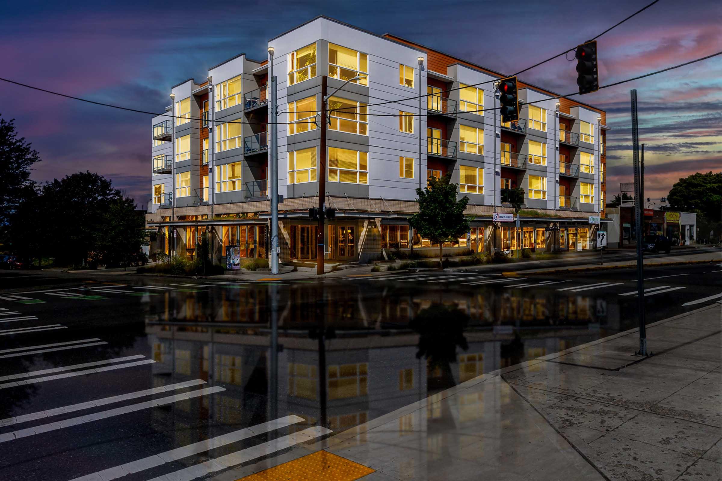 A modern multi-story building with many windows illuminated at dusk, reflecting in a wet street. Traffic lights hang above, and the scene includes crosswalks and nearby trees. The sky features vibrant colors as the sun sets.
