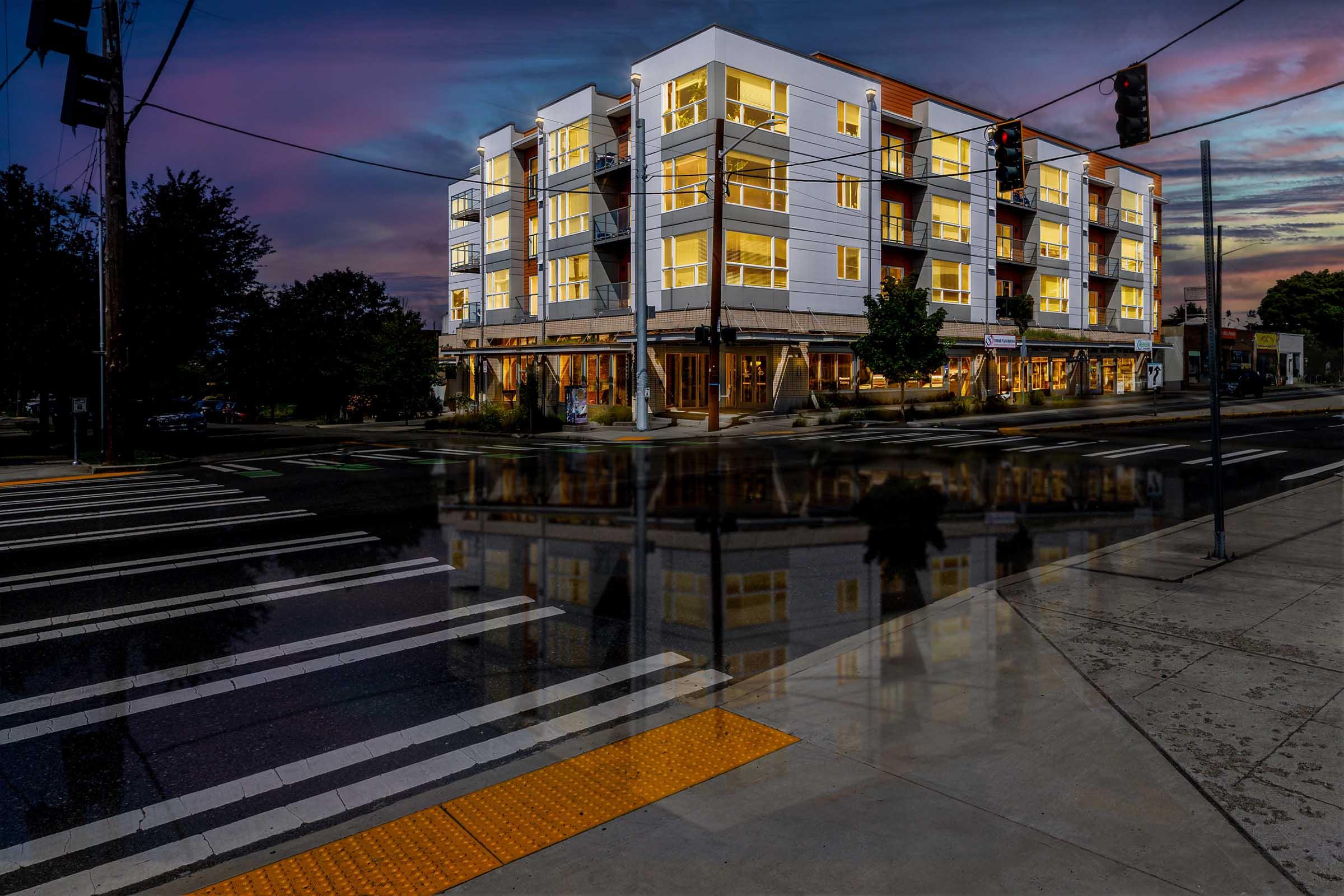 A modern apartment building with illuminated windows, situated at a street corner. The scene is captured during twilight, showcasing a colorful sky. Reflections of the building and traffic lights are visible on a wet road, enhancing the atmospheric feel of the urban setting.