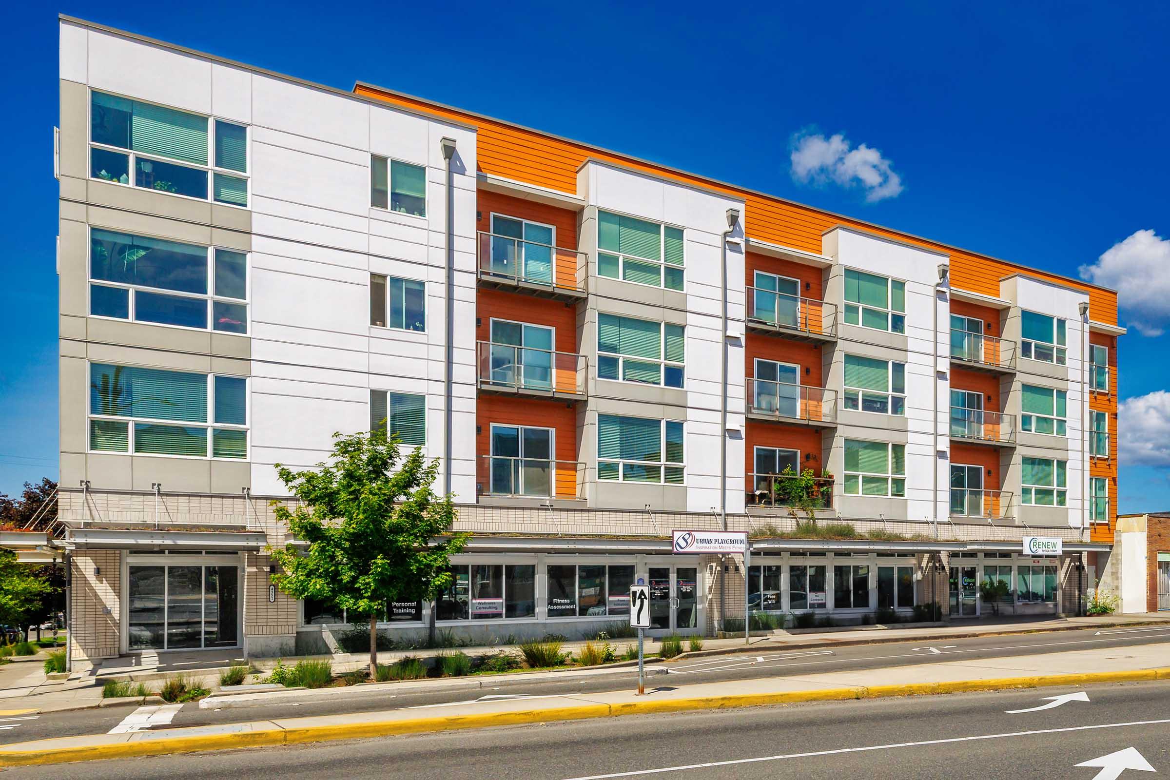 Modern multi-story apartment building with a mix of white and orange exterior. The building features balconies on each unit, large windows, and ground-level storefronts. The surrounding area has trees and a clear blue sky, creating a welcoming urban environment.