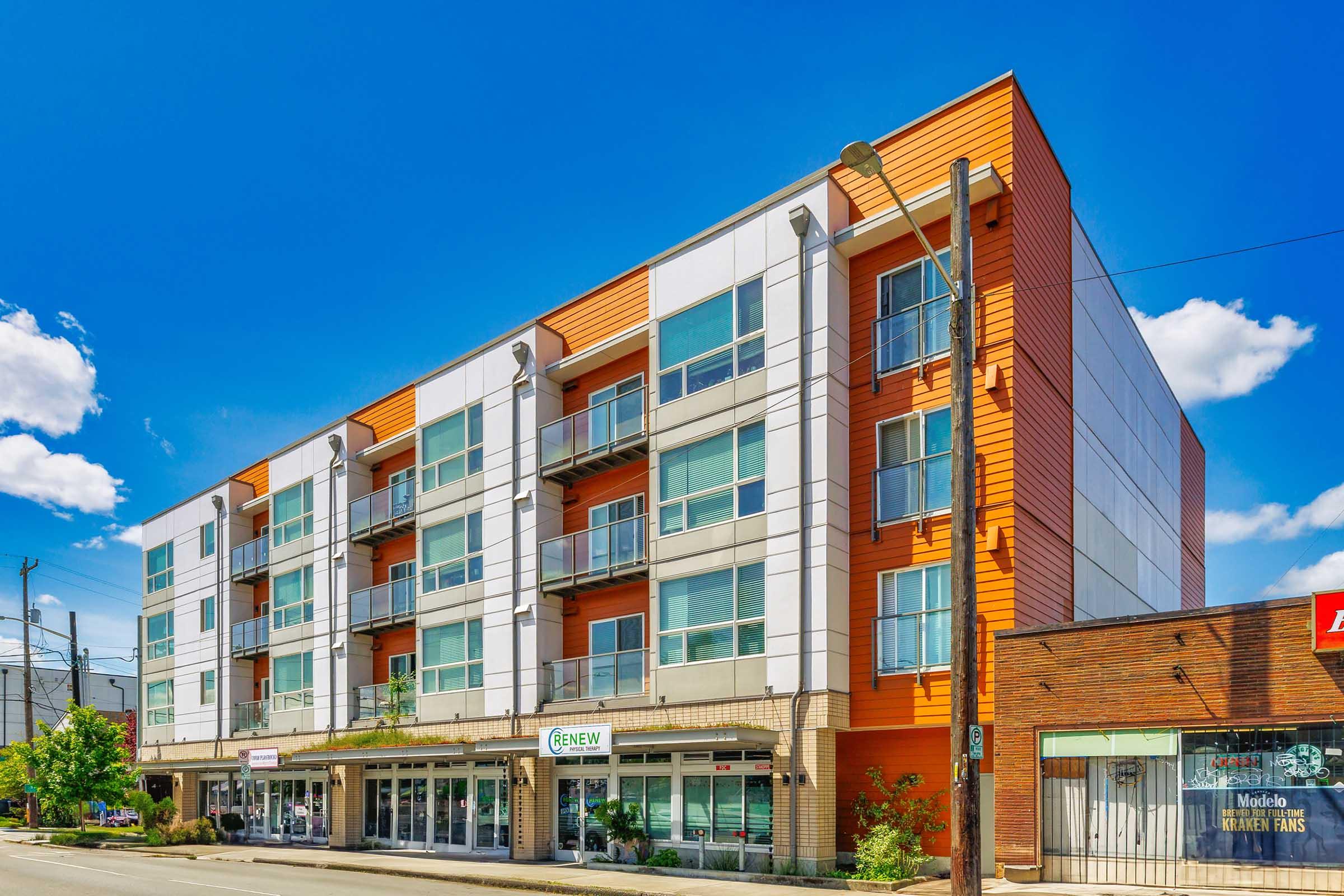A modern four-story building with a mix of orange and white facades, featuring large windows and balconies. The ground floor includes storefronts with signage, and the surrounding area has trees and a clear blue sky.
