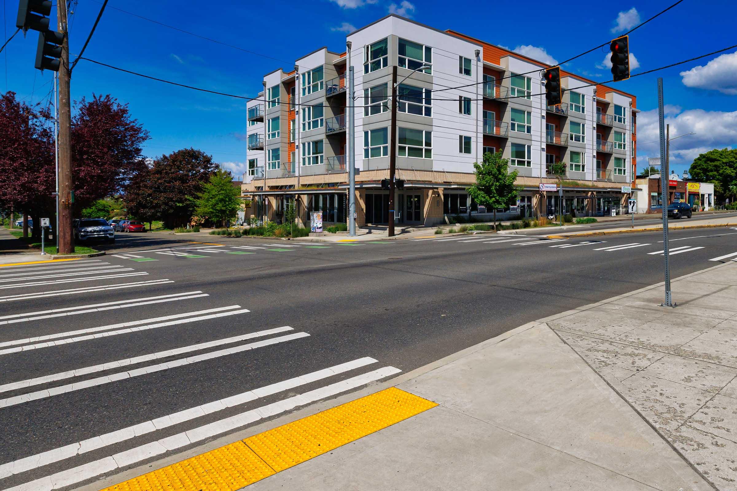 A street view of a modern building with multiple floors on the corner of an intersection. The building features large windows and balconies, surrounded by trees and small shrubs. Pedestrian crosswalks are visible on the road, with a traffic light and sidewalk on the right. Bright blue sky with some clouds.
